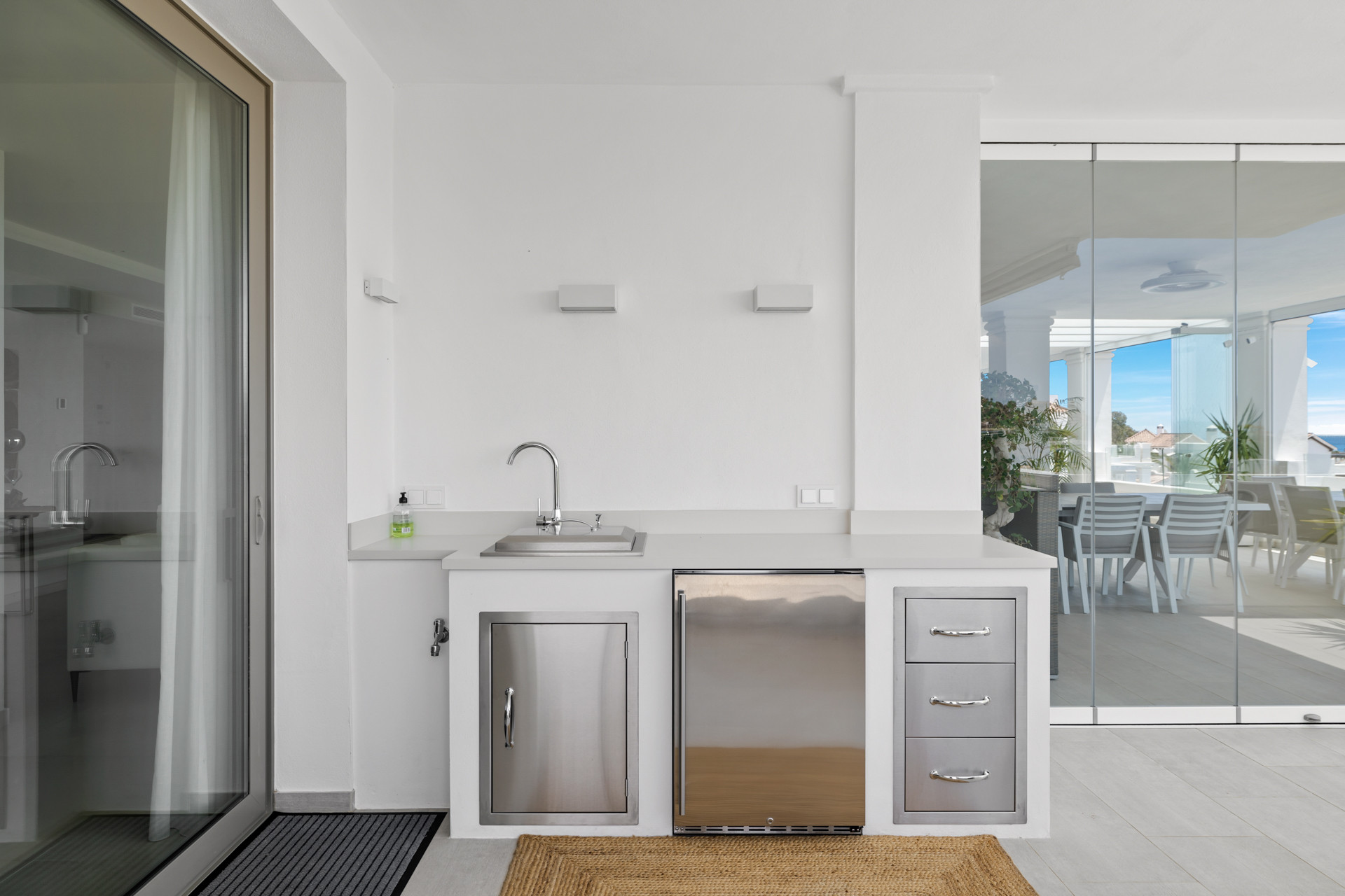 Outdoor kitchen counter with sink, under-counter refrigerator, and drawers next to a sliding glass door.