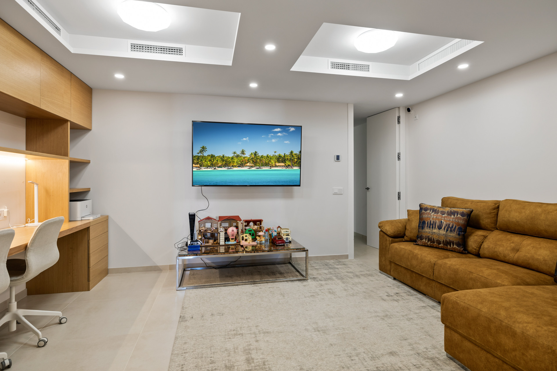 Living room with a large tan sectional sofa, wall-mounted TV, and a wooden desk area with a chair.