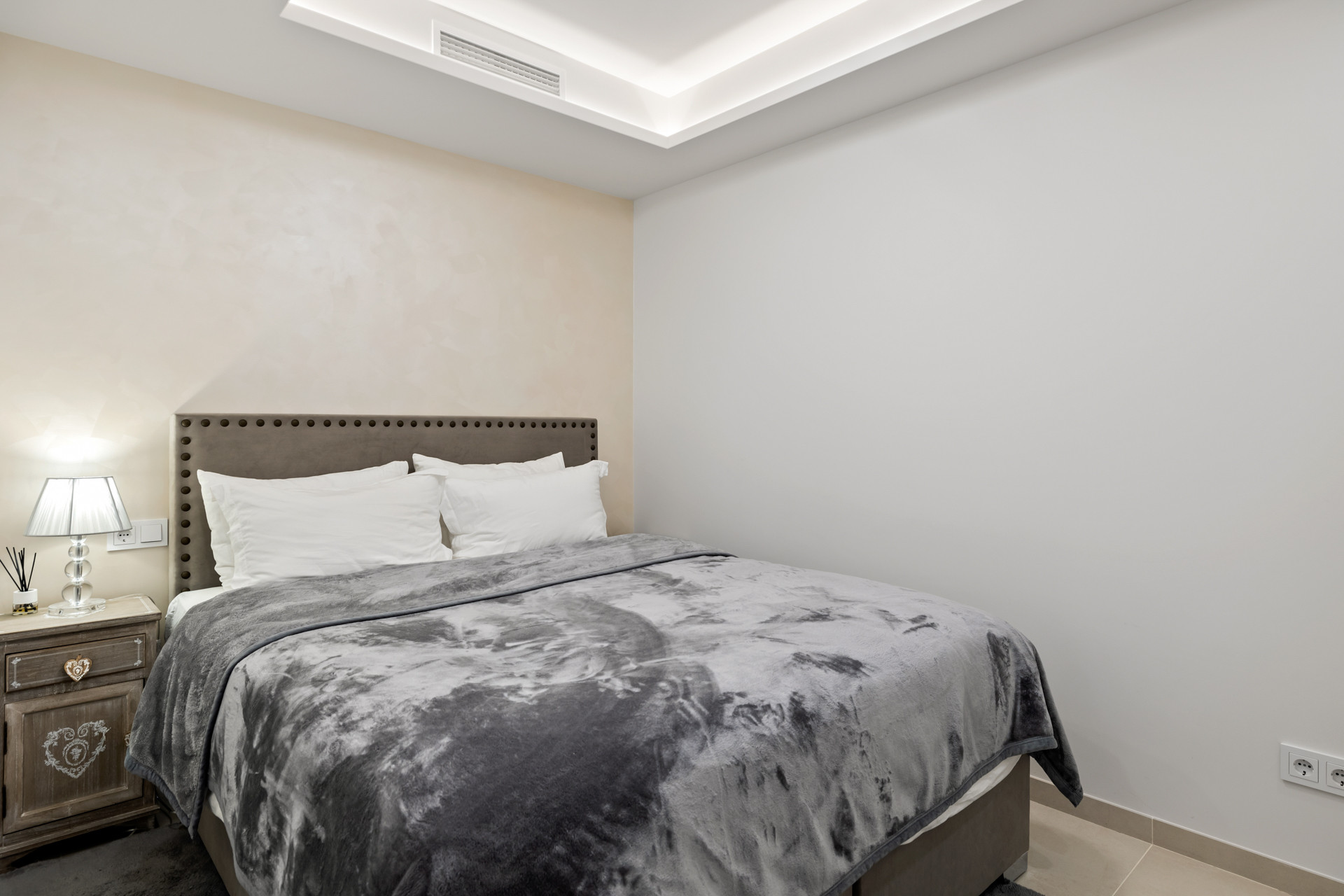 Bedroom with a gray upholstered headboard, white and textured beige walls, and recessed lighting.