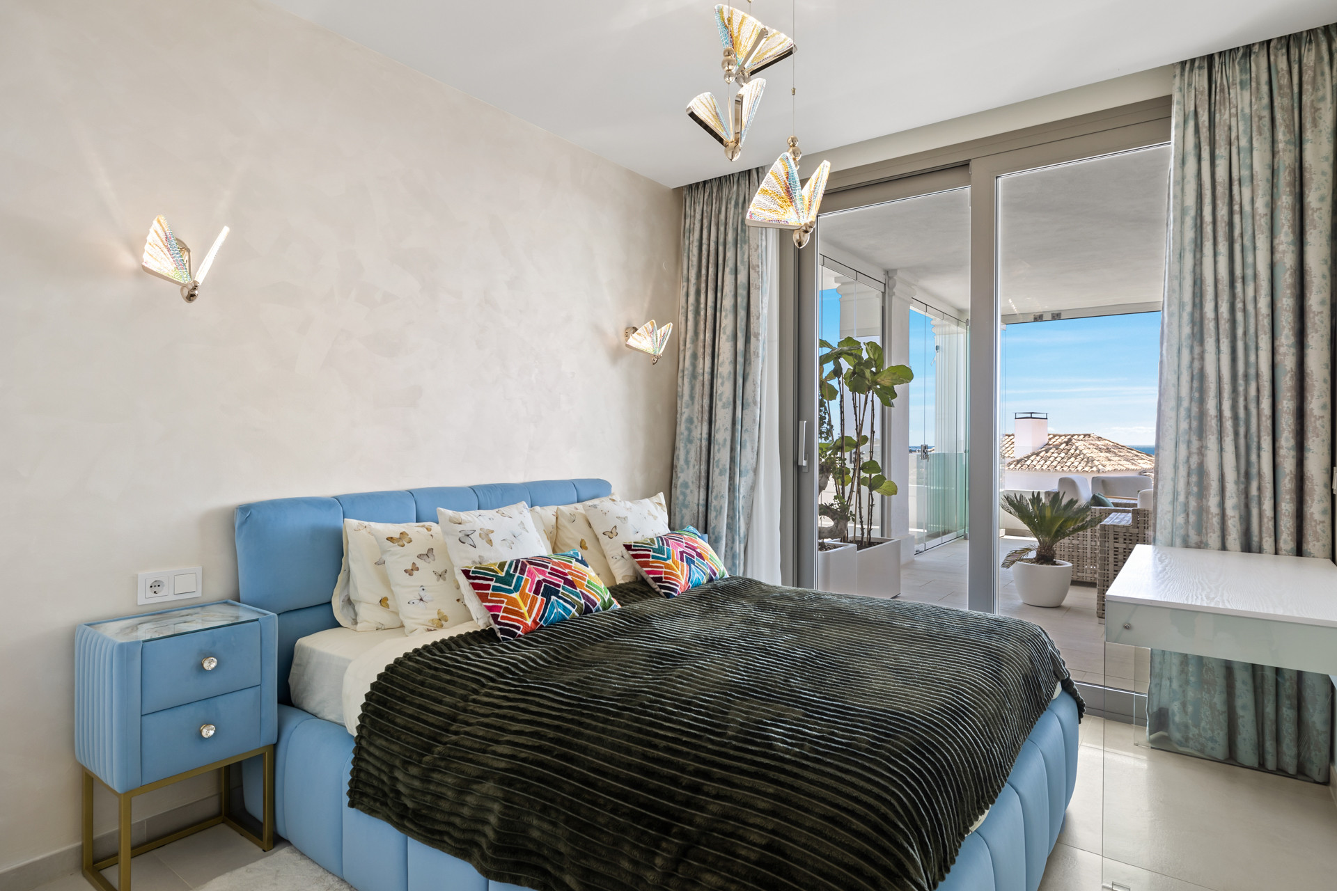 Bedroom with a blue bed, colorful pillows, and butterfly shaped wall sconces, leading to a balcony.