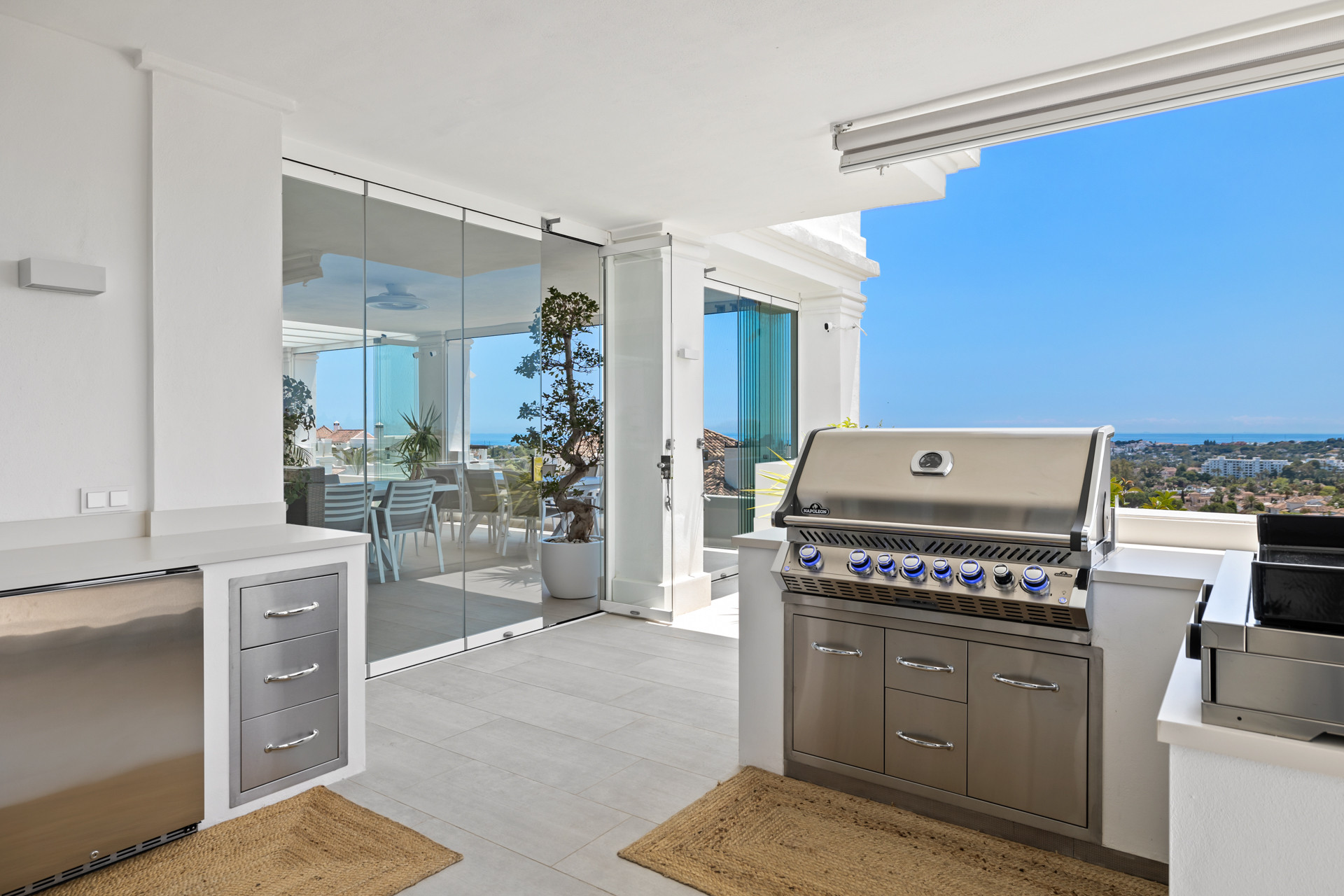 Outdoor kitchen with stainless steel grill and appliances, glass doors, and a distant sea view.