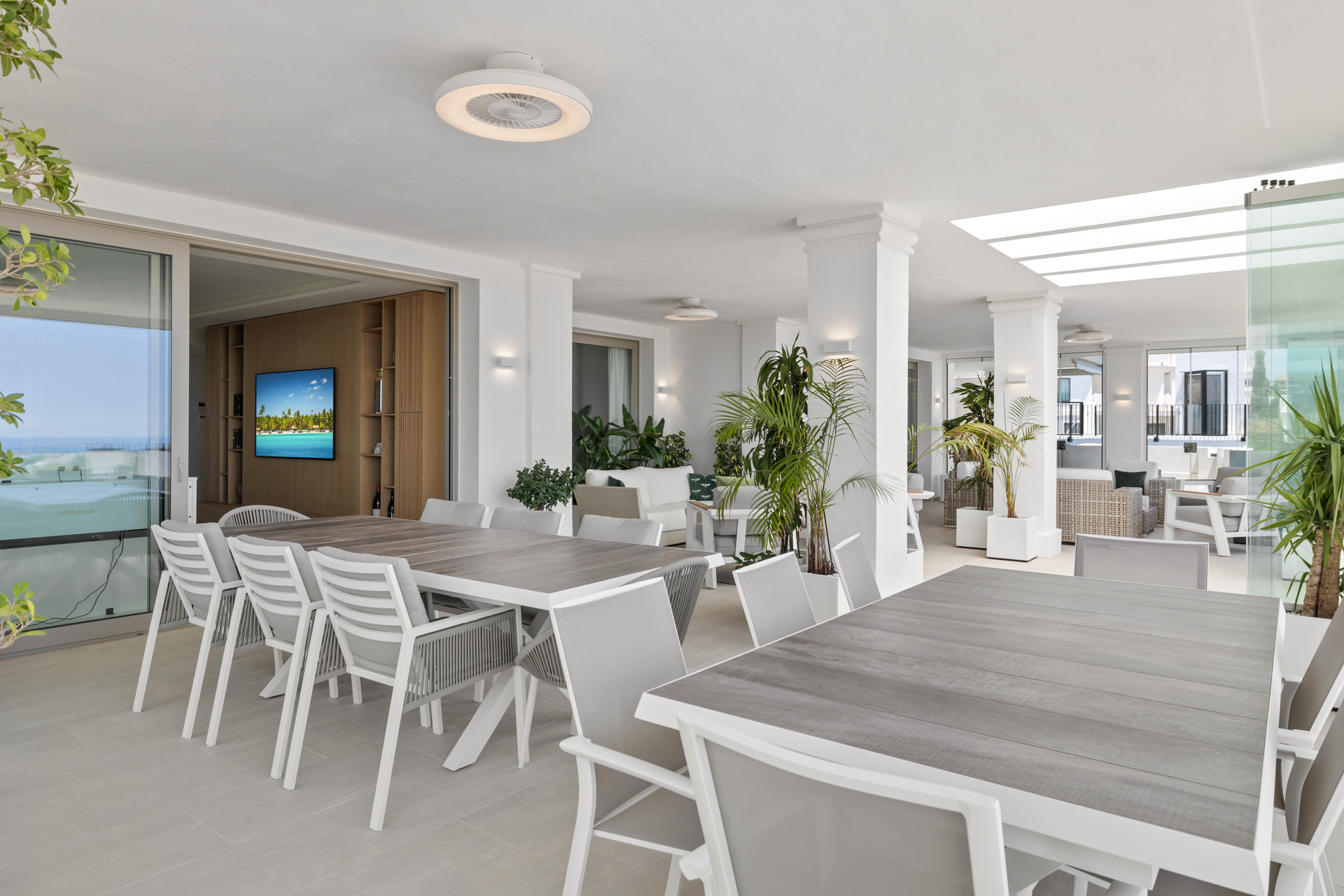 Covered outdoor dining area with multiple tables, plants, and a partial view into an interior room with a TV.