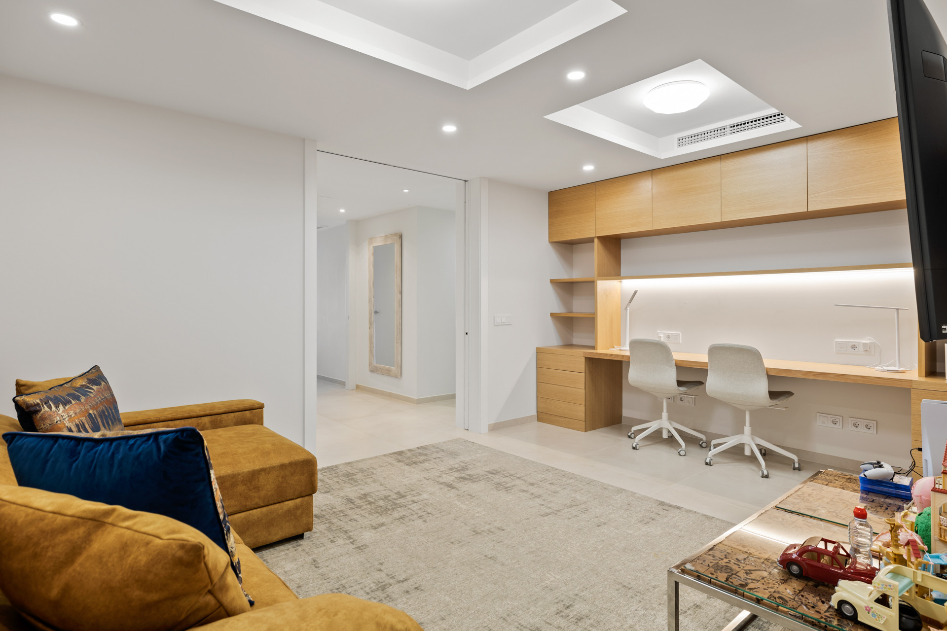 Living room with a tan sectional sofa, a built-in wooden desk with two chairs, and a hallway entrance.