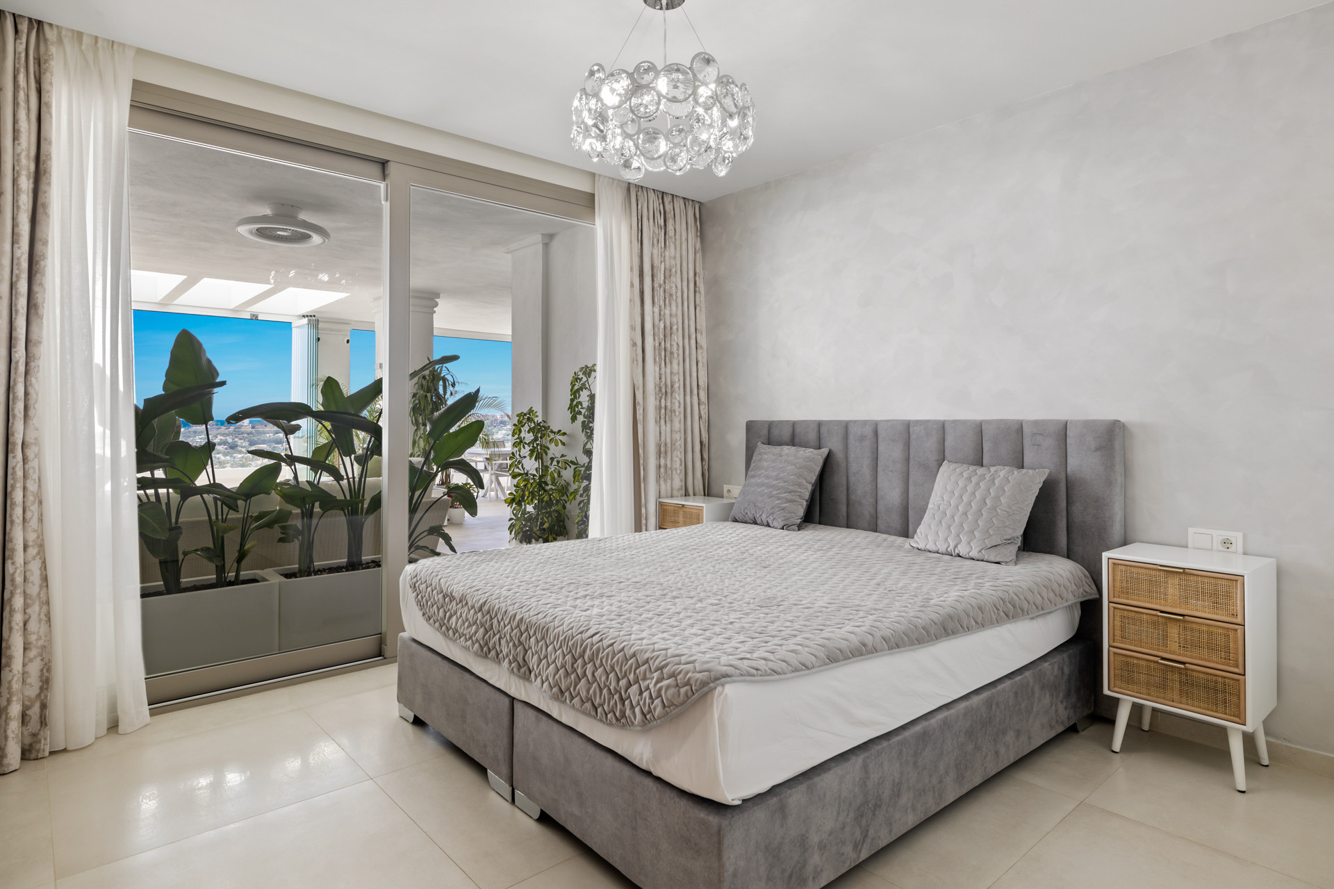 Bedroom with a gray bed, crystal chandelier, and sliding glass doors to a plant-filled balcony with views.
