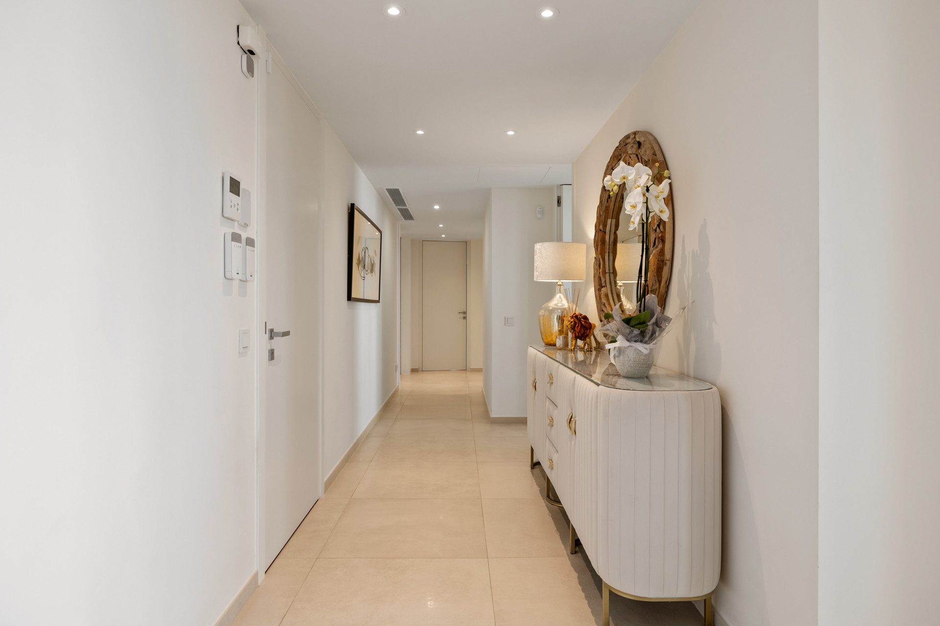 Long white hallway with light tile flooring, an ornate console table with a mirror, and recessed lighting.