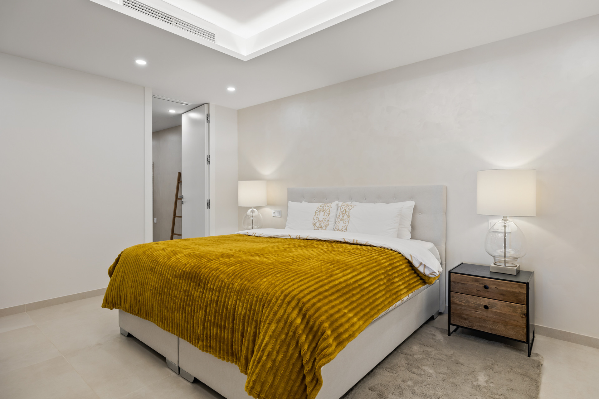 Bedroom with a large bed, yellow comforter, two nightstands with lamps, and a partial view of a bathroom.
