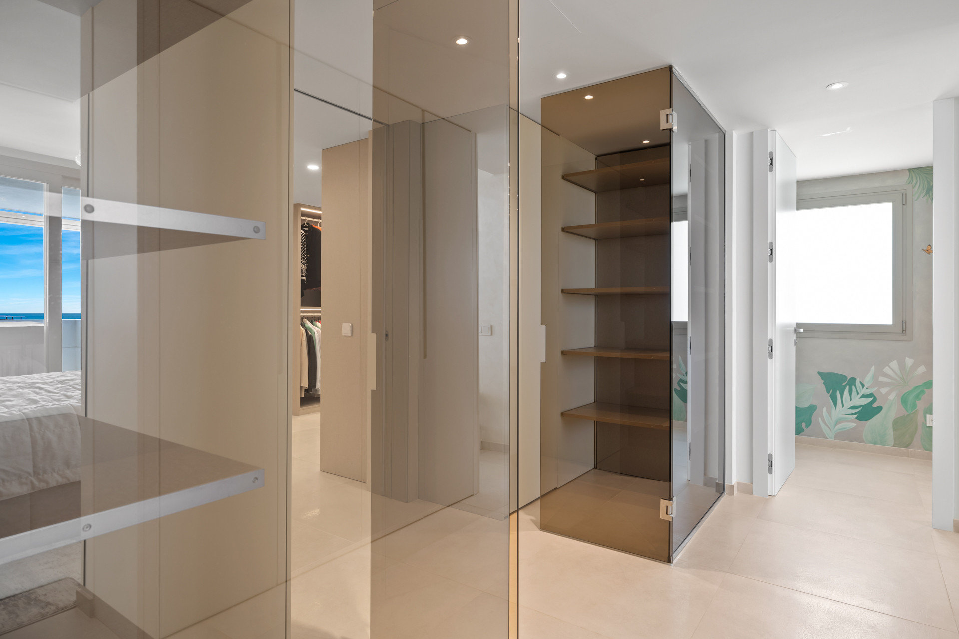 Walk-in closet area with glass and light wood shelving, leading to a hallway and another room.