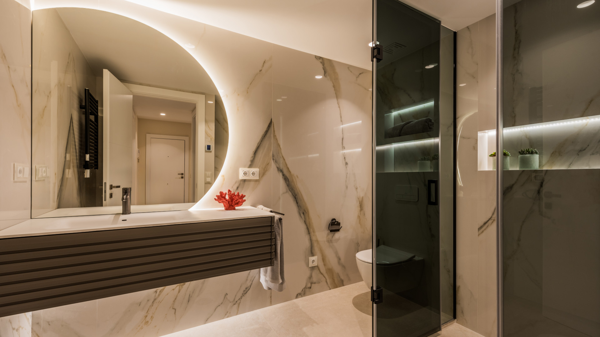Modern bathroom with illuminated mirror, floating vanity, toilet, and glass shower enclosure.