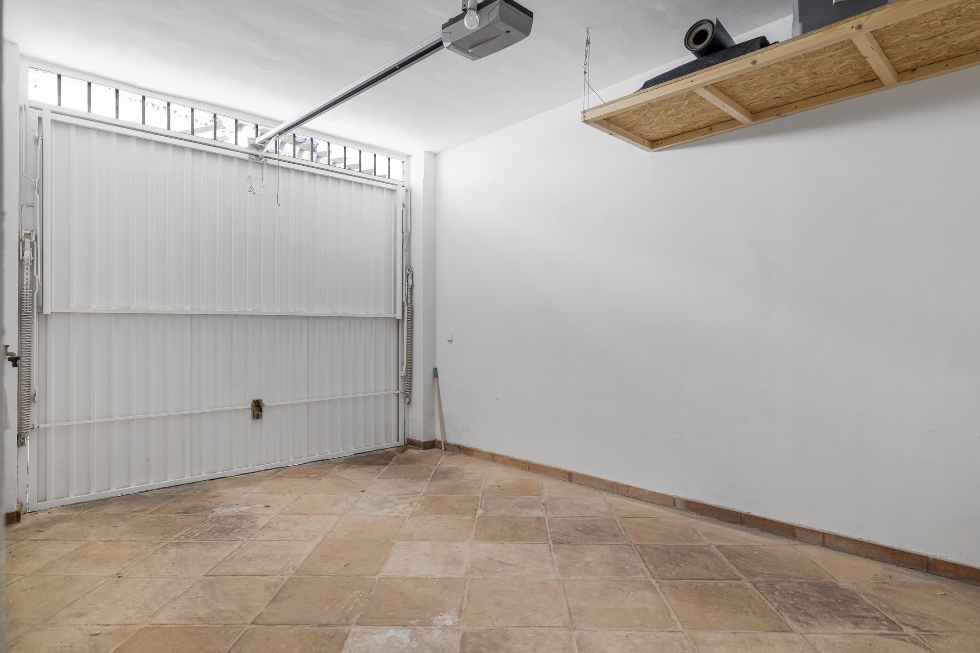 Empty garage with a white sectional door, tiled floor, white walls, and an overhead wooden storage shelf.