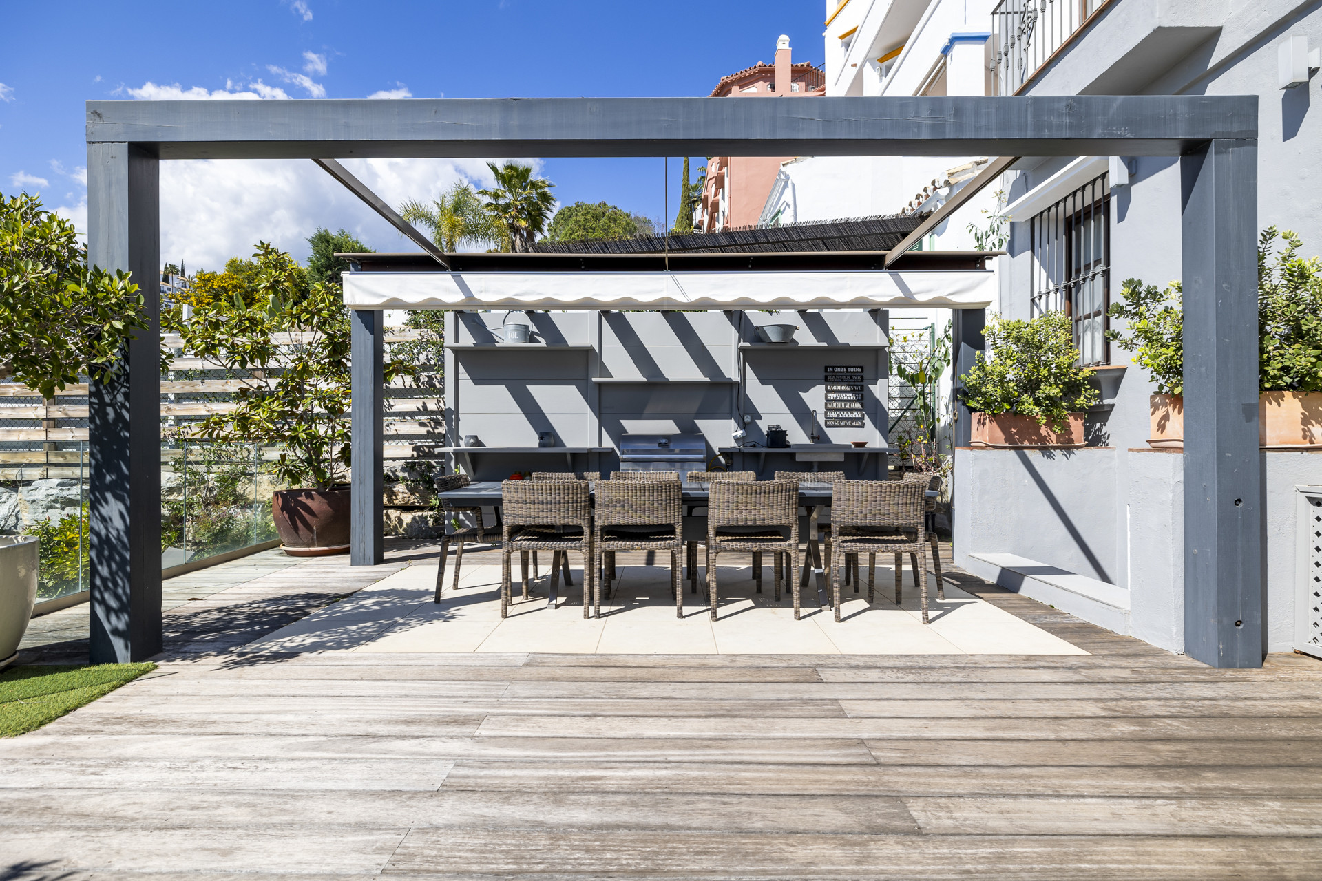 Outdoor kitchen and dining area under a grey pergola, with a long table and six chairs on a light patio.