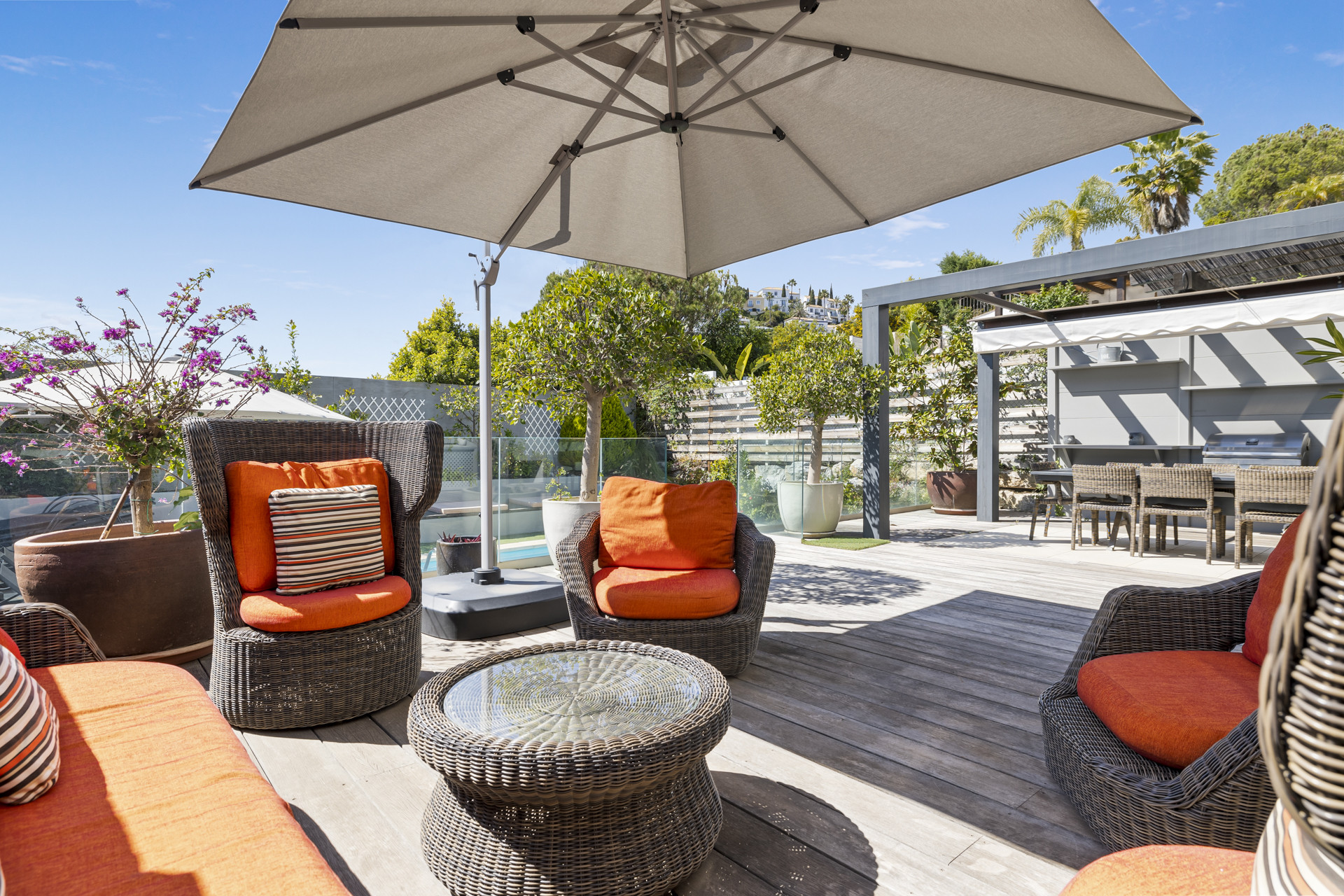 Outdoor lounge area with wicker chairs, orange cushions, a glass table, and a large umbrella on a wooden deck.