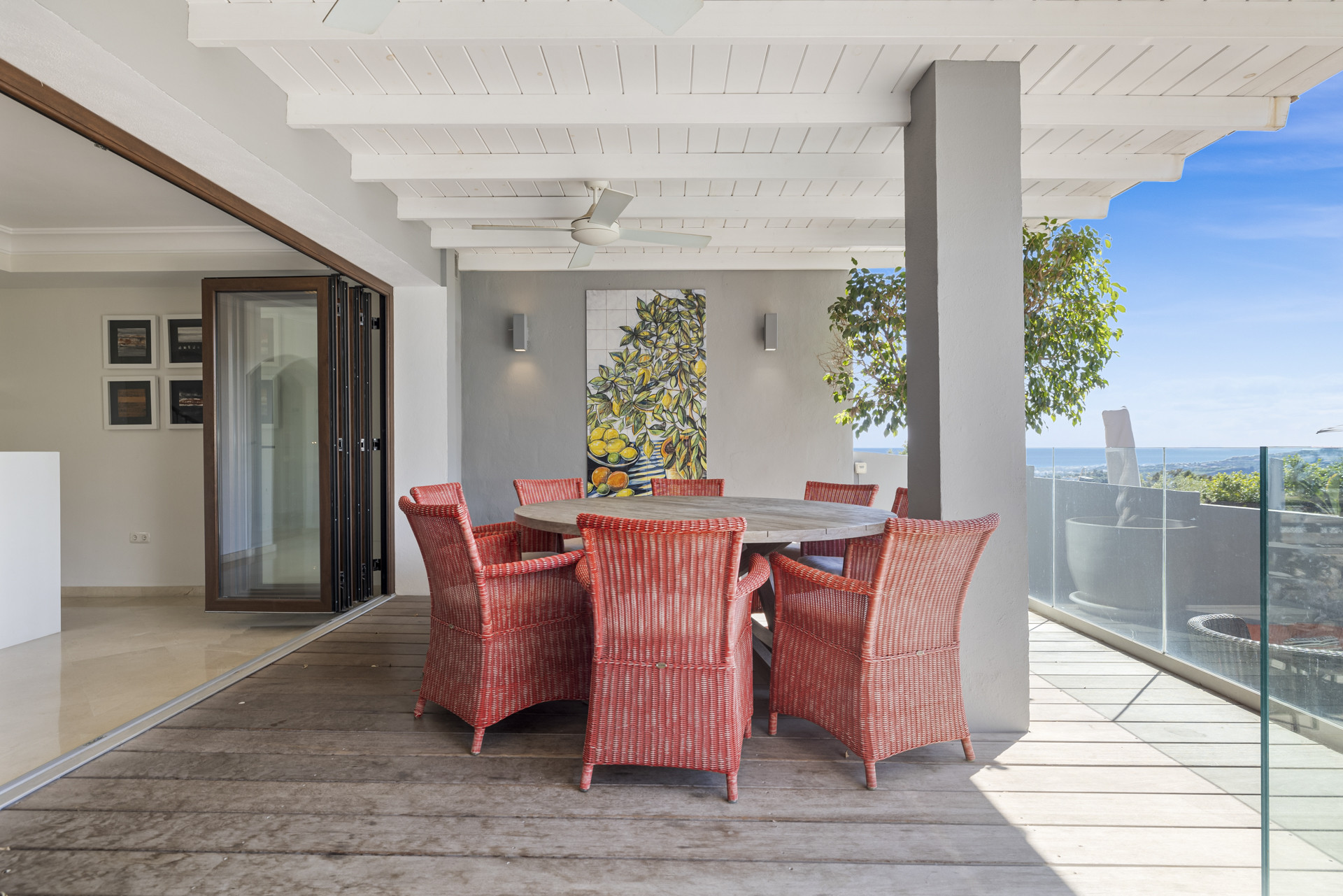 Covered patio with a wooden table, red wicker chairs, and a glass railing with a distant sea view.