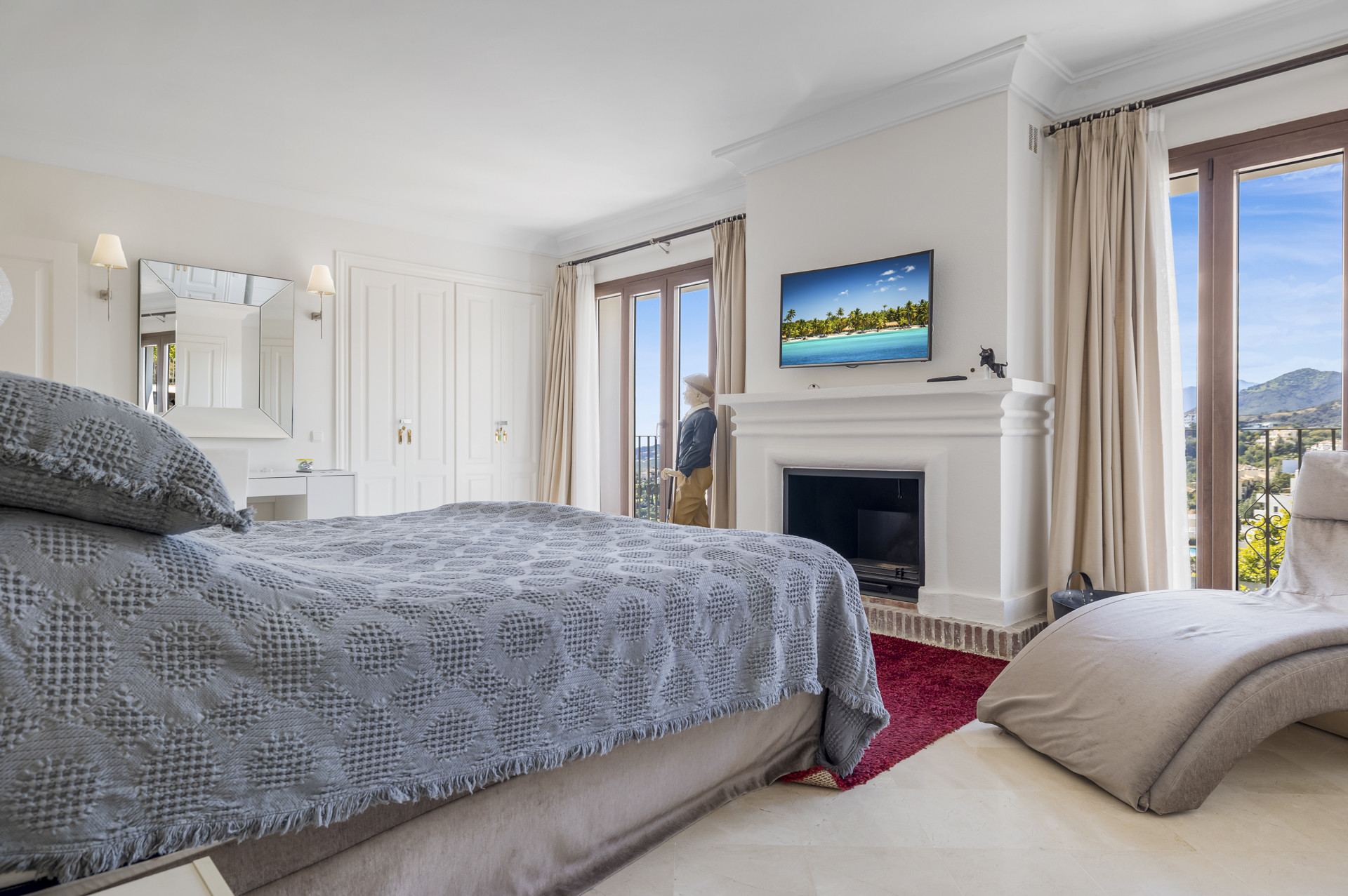 Bedroom with a bed in foreground, fireplace, TV, and a recliner chair near balcony doors.