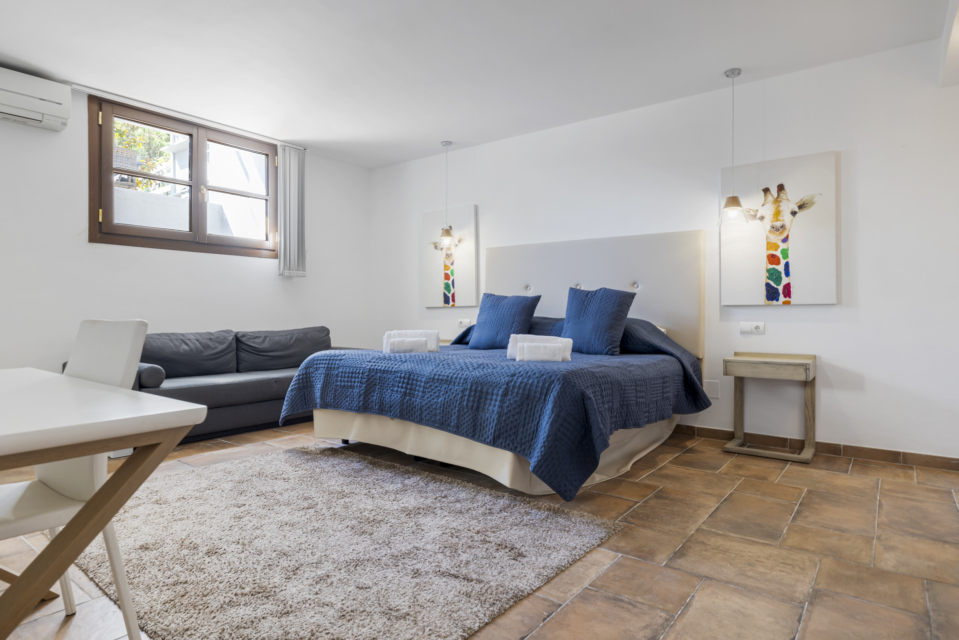 Bedroom with a large blue bed, two giraffe artworks, a gray sofa, and a white table.
