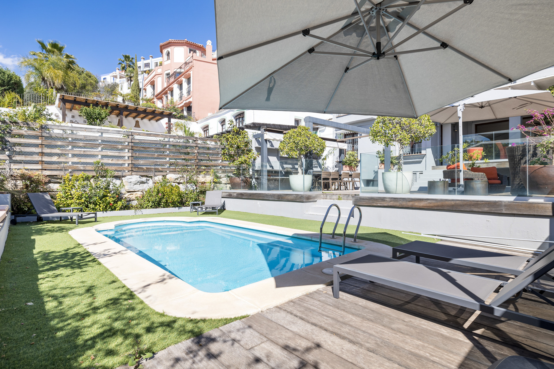 Swimming pool with a ladder next to artificial grass, leading to a wooden deck and townhouse exterior in sunlight.