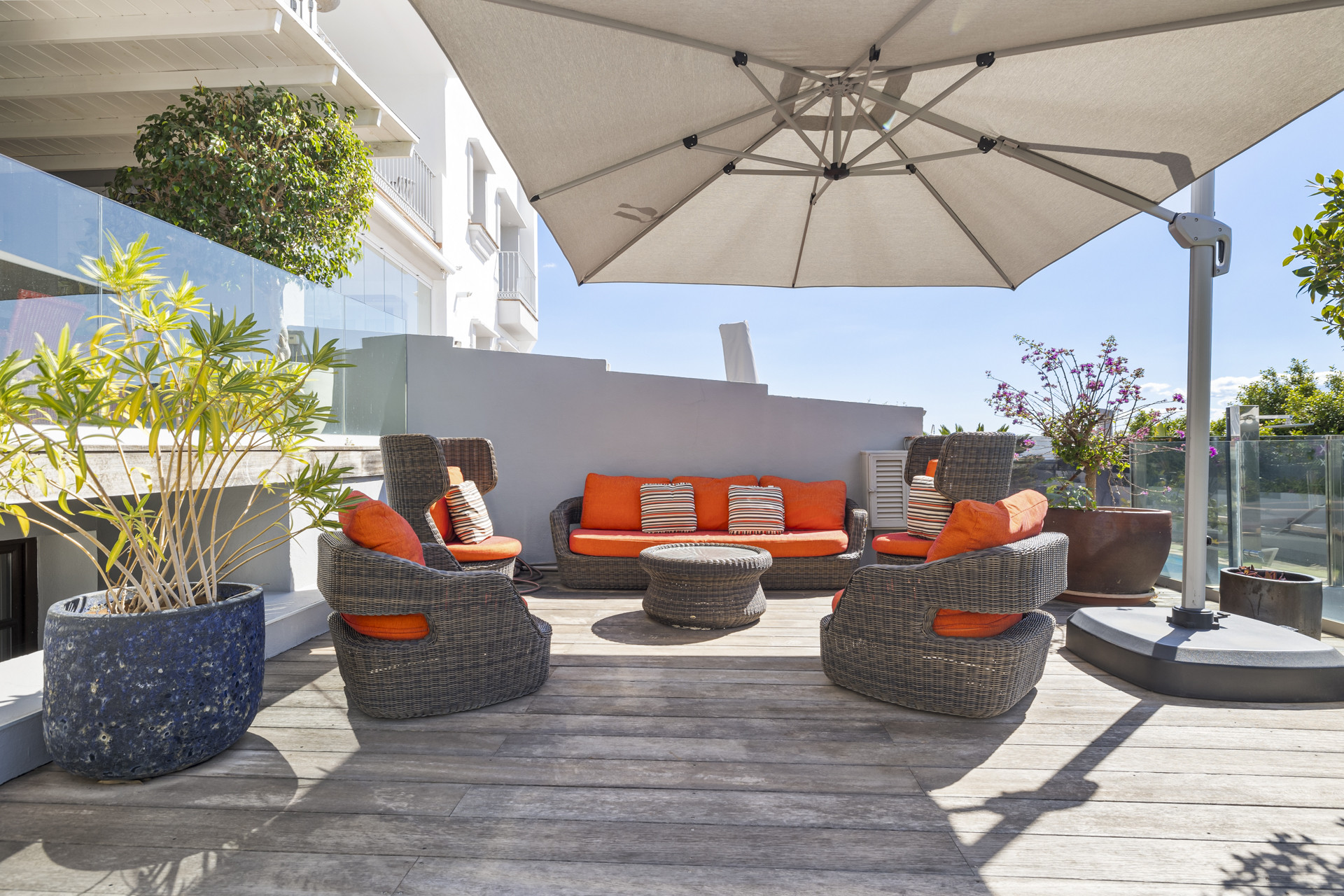 Outdoor terrace with wicker furniture, orange cushions, potted plants, and a large open umbrella.
