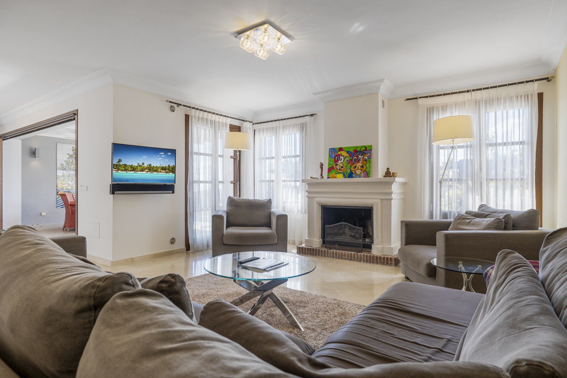 View of a living room with fireplace, gray seating, glass coffee table, and large windows.
