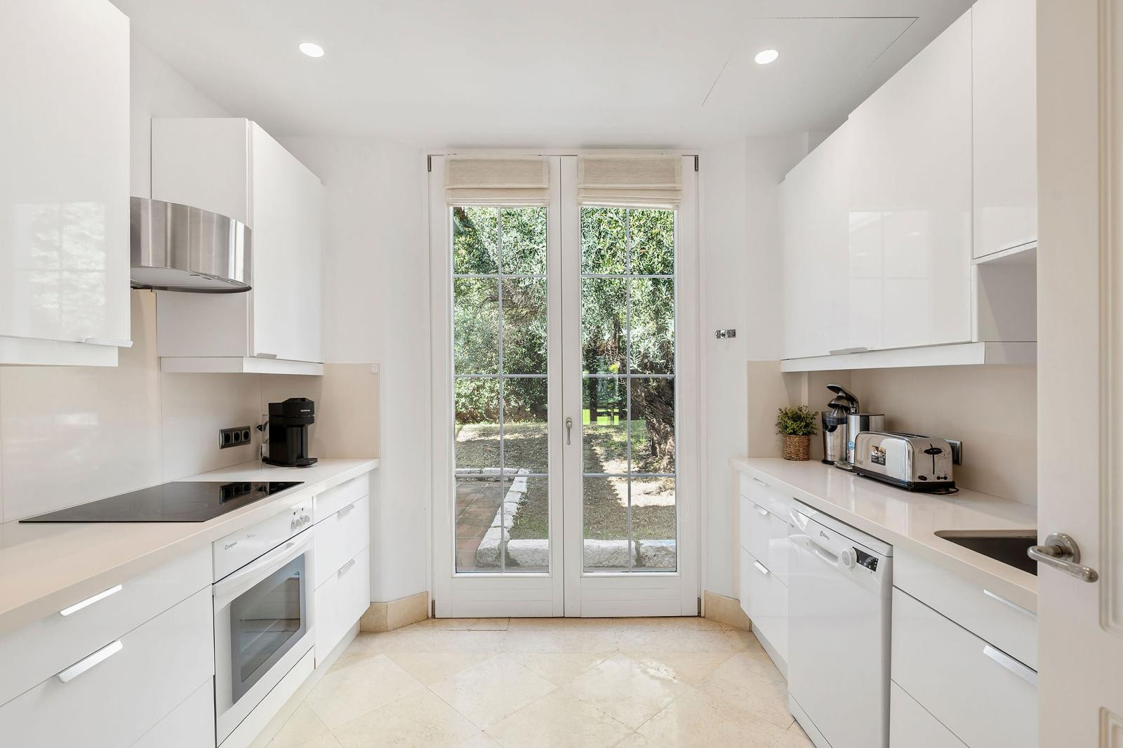 Narrow white kitchen with a coffee machine, toaster, and glass doors opening to a garden view.