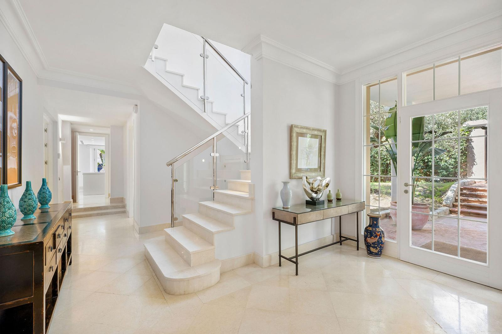 Bright white entryway with a modern marble staircase, glass railing, console table, and glass door to garden.