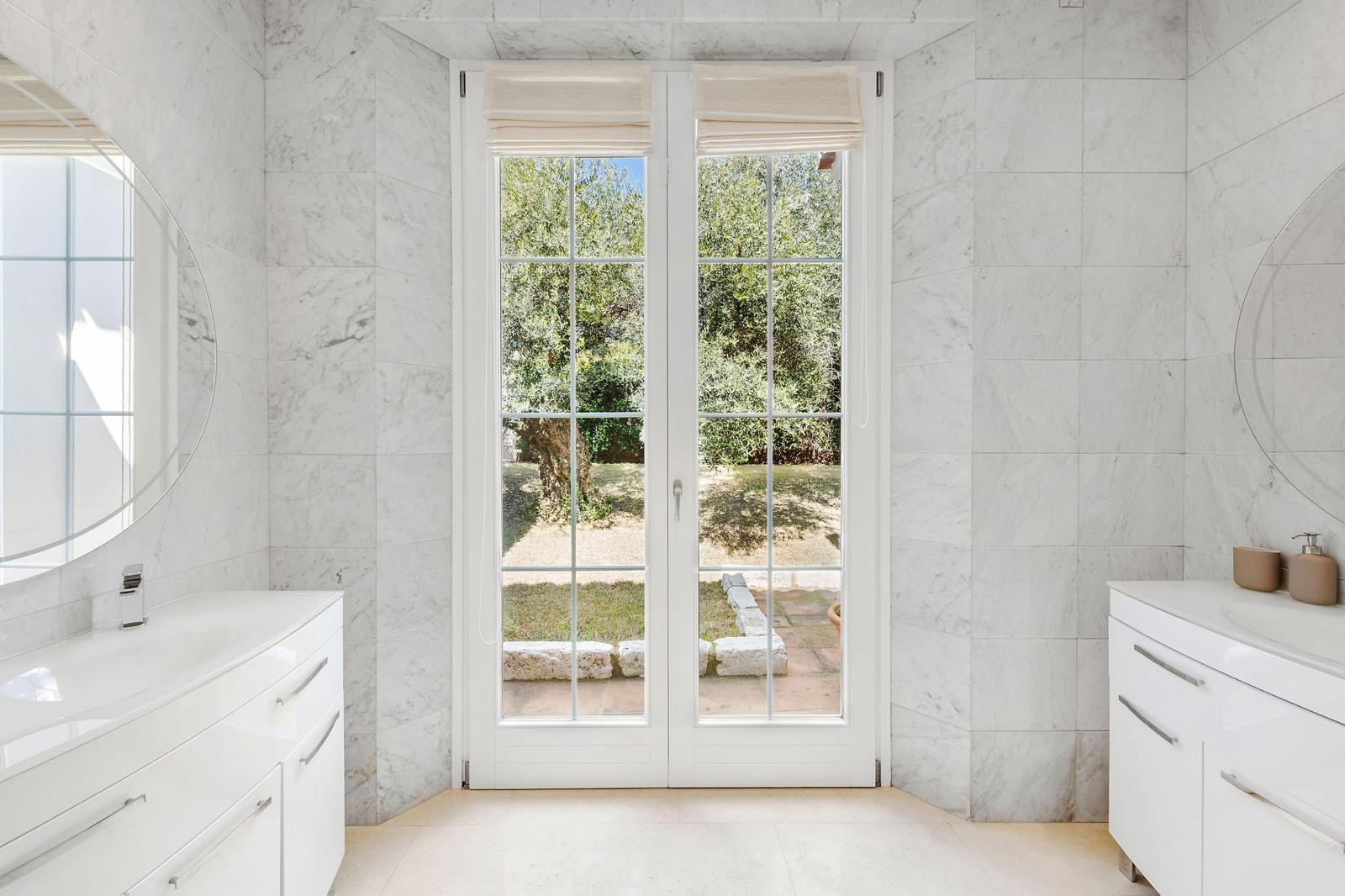 Bathroom with white marble walls, two vanities, and French doors opening to a garden.
