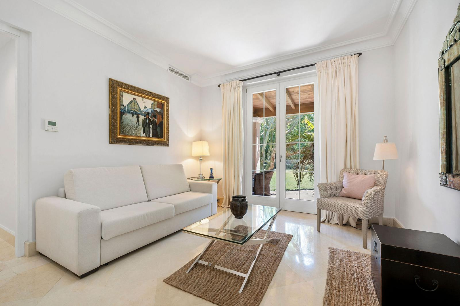 Living room with a cream sofa, glass coffee table, armchair, artwork, and French doors to a garden.