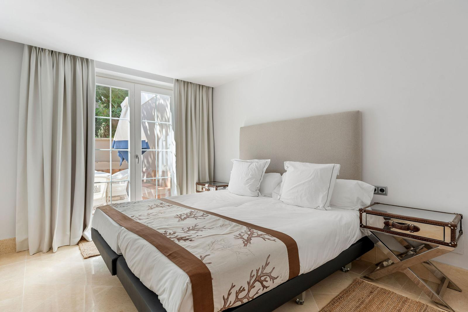 Bedroom with a white bed, beige headboard, mirrored nightstands, and French doors leading to an outdoor area.