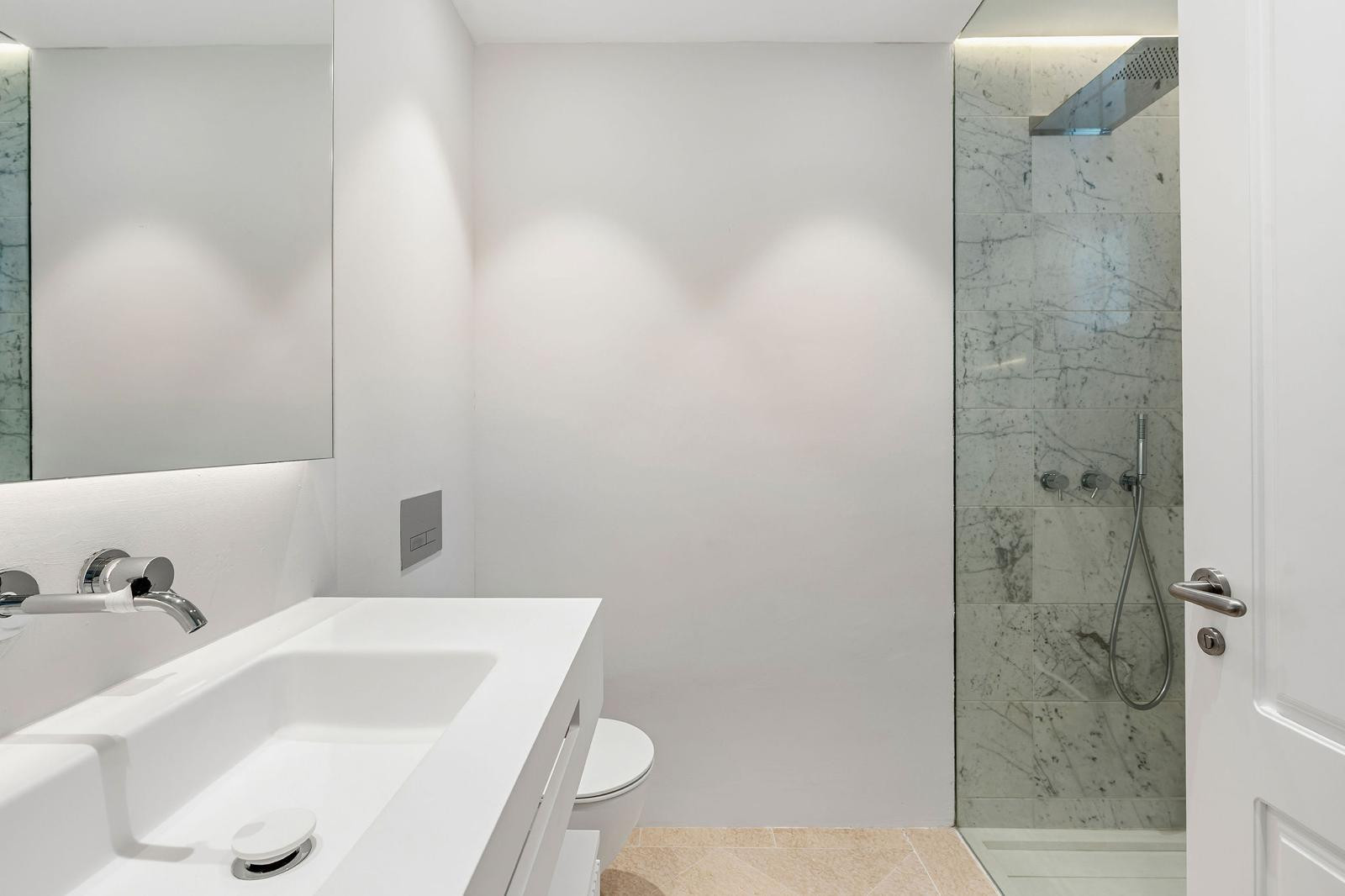 White bathroom with a rectangular sink, wall-mounted faucet, toilet, and a walk-in shower with marble tiles.