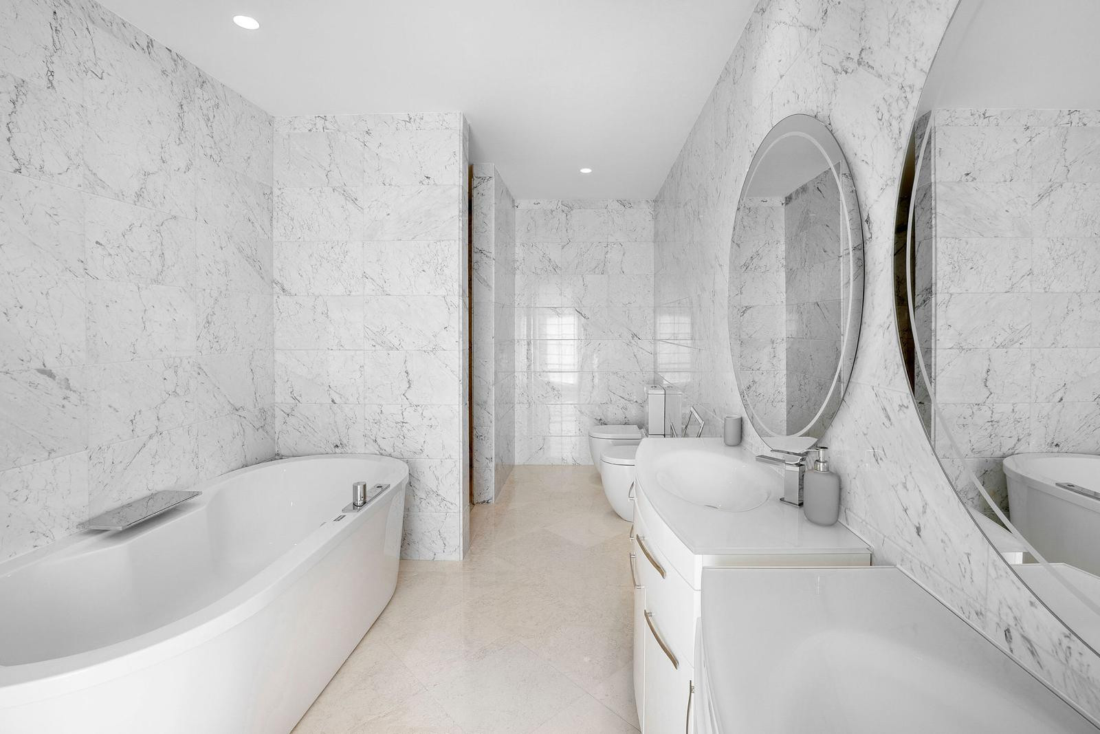 White marble bathroom with a freestanding tub, double vanity, two oval mirrors, and a toilet.