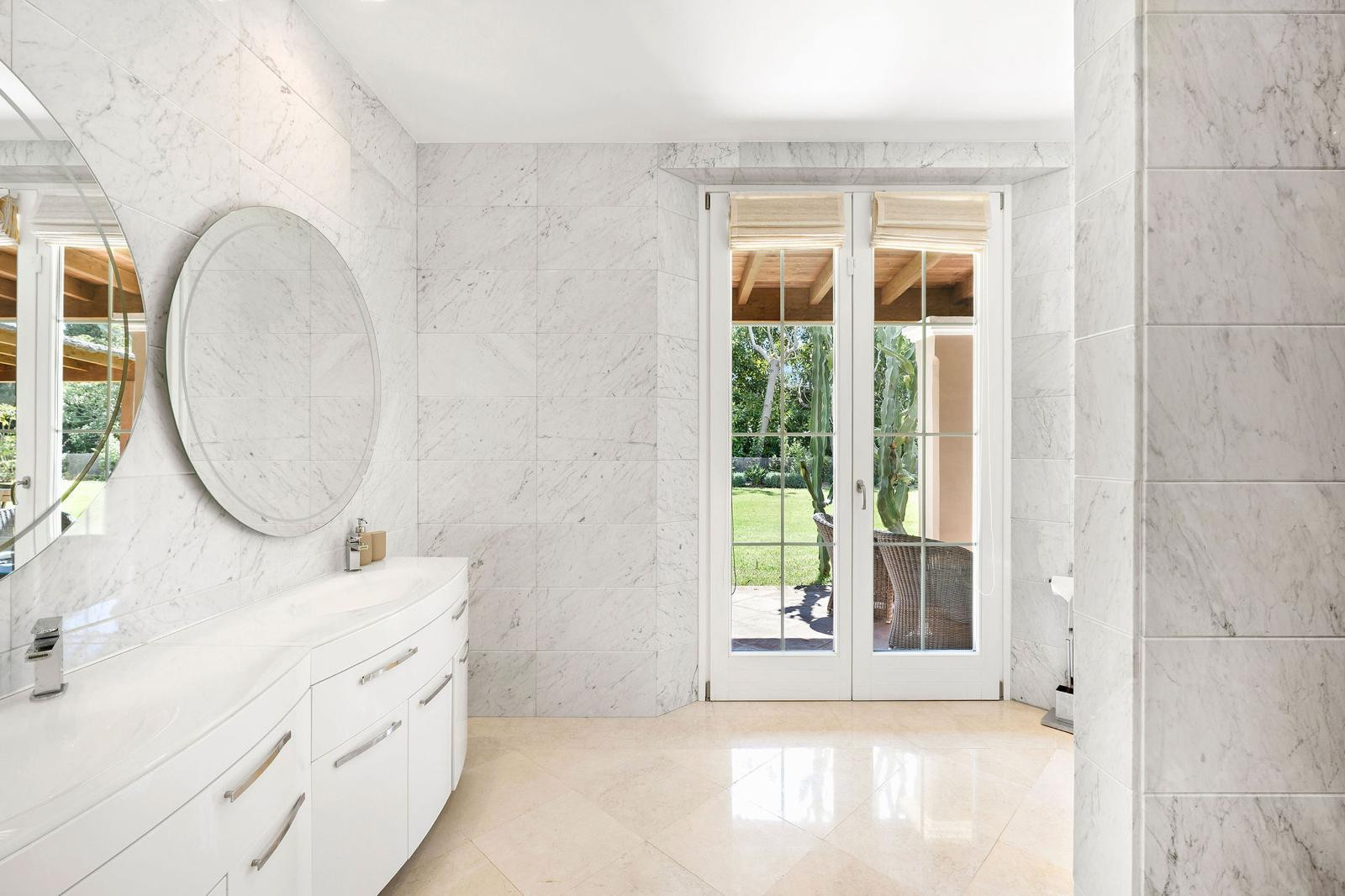 Bathroom with a white curved double vanity, two round mirrors, and French doors opening to a lush garden.