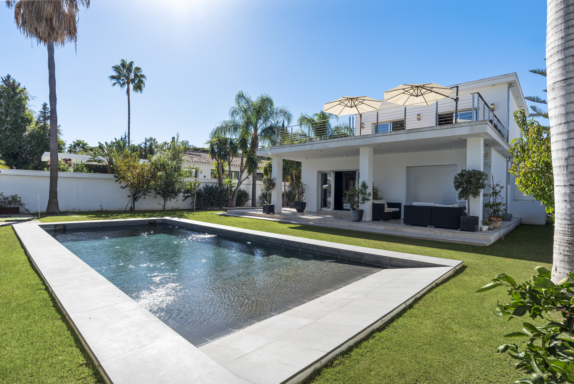 Rear view of a white villa with a swimming pool, grassy lawn, large patio, and rooftop terrace.