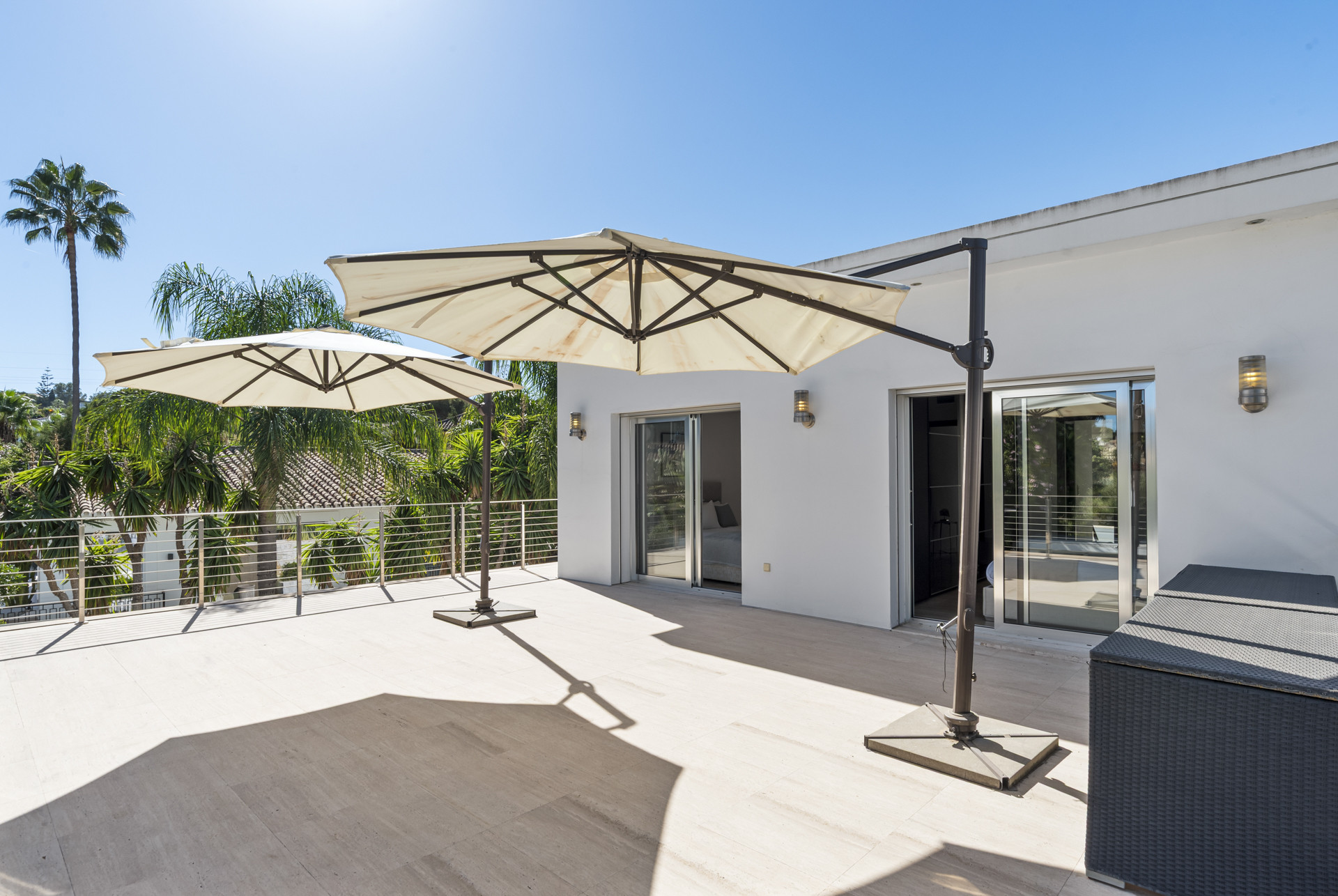 Wide outdoor terrace with two large beige umbrellas, white walls, glass sliding doors, and distant palm trees.
