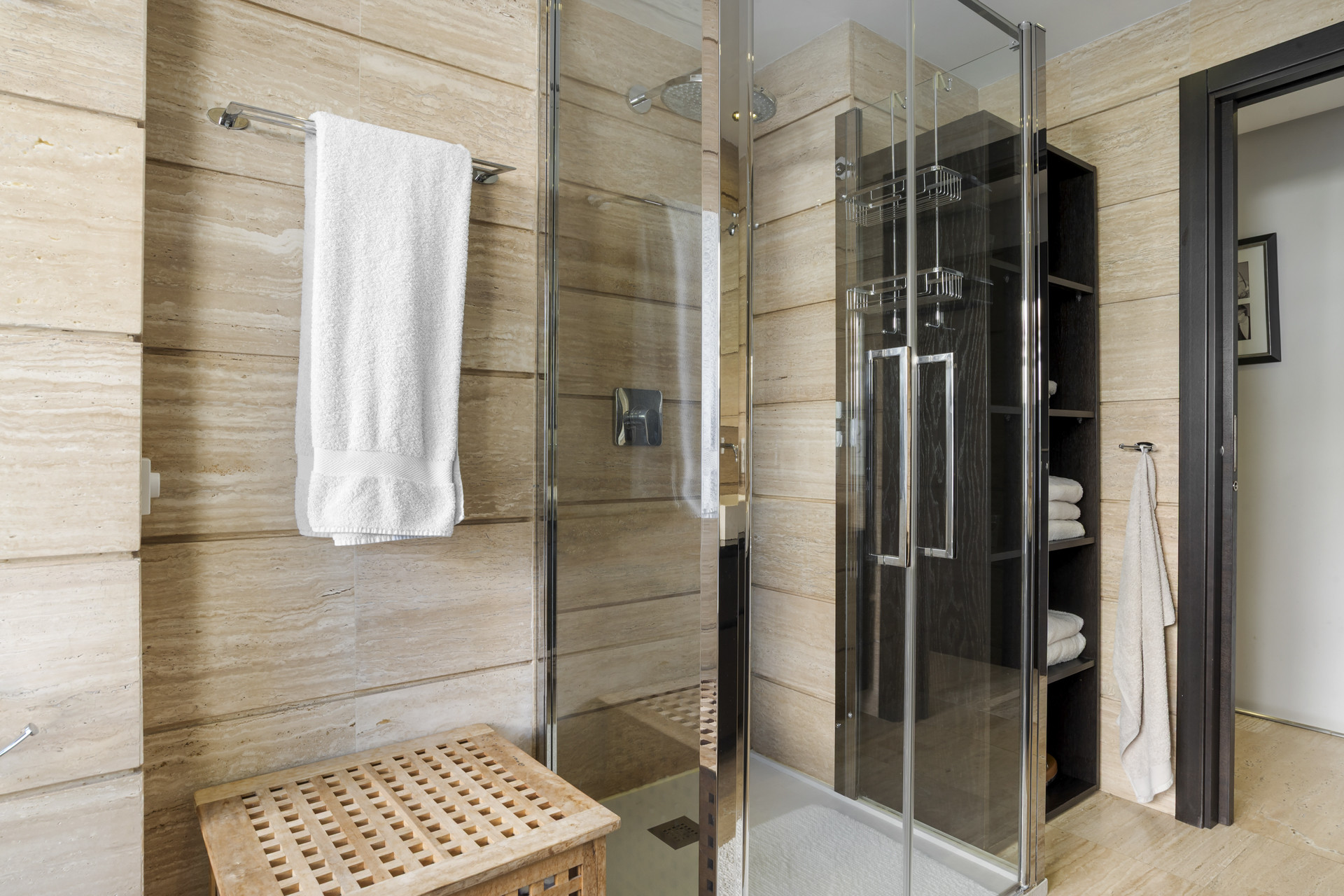 Walk-in shower with glass doors, a wooden slatted bench, and a towel rail in a tiled bathroom.