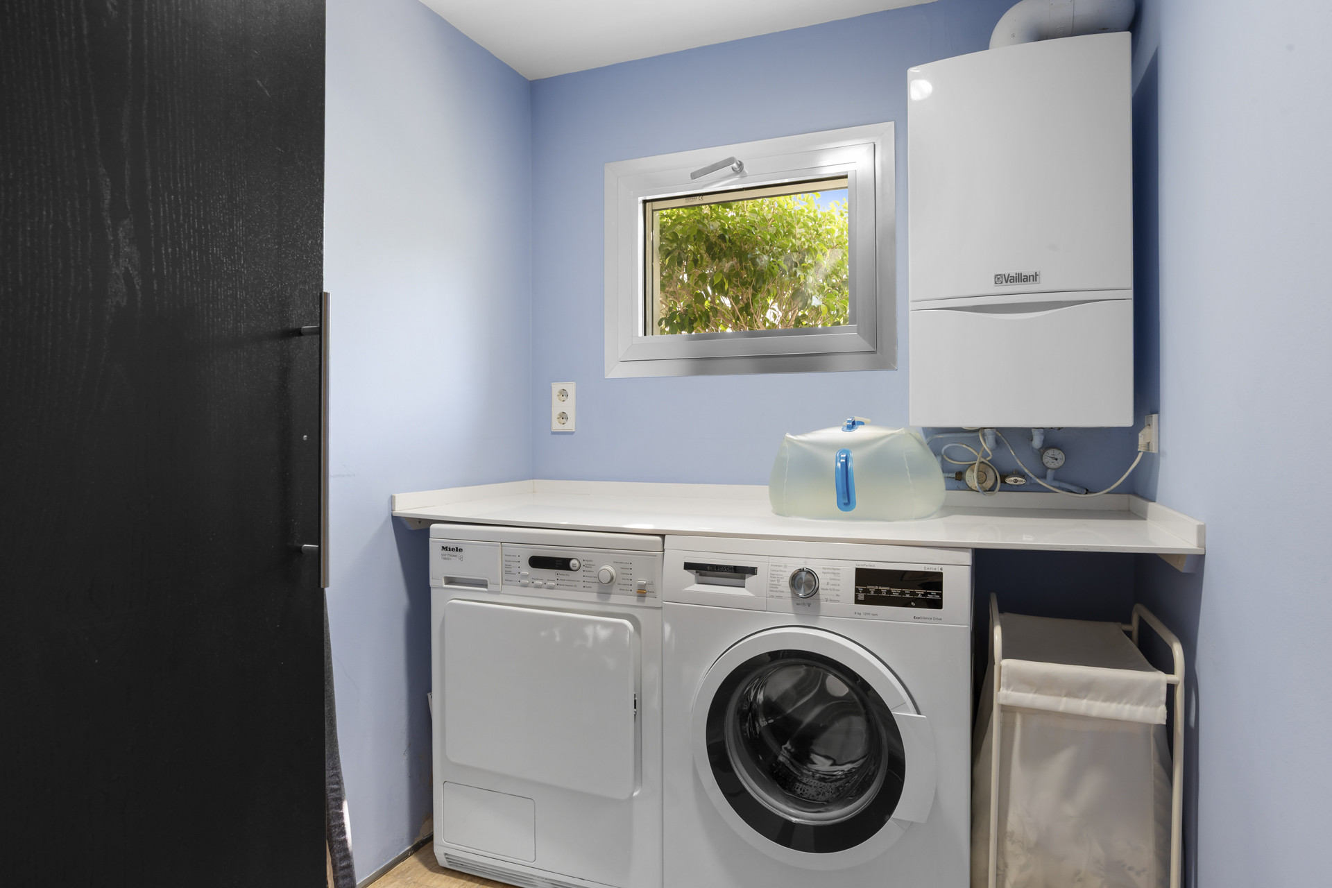 A laundry room with a Miele washer and dryer, blue walls, a water heater, and a small window.
