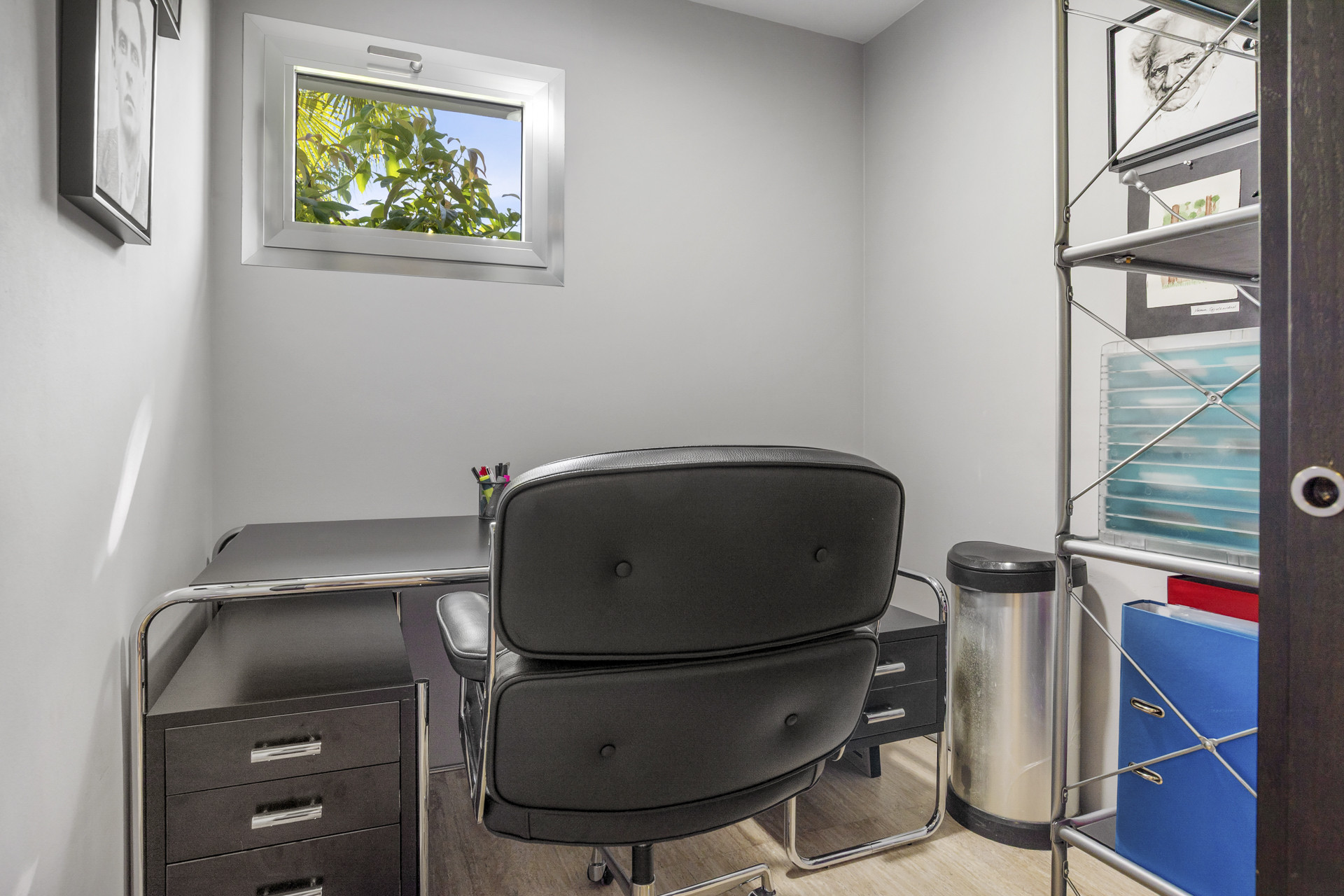 Small home office with a black desk, rolling chair, and a small window showing greenery.