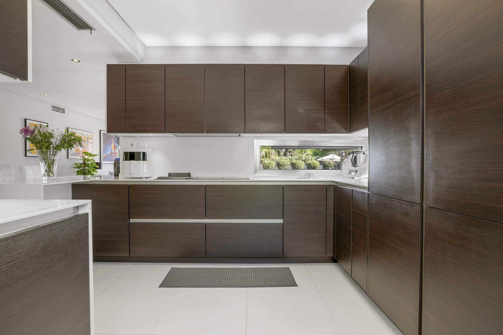 Modern kitchen with dark wood cabinetry, white countertops, and a long horizontal window above the sink.
