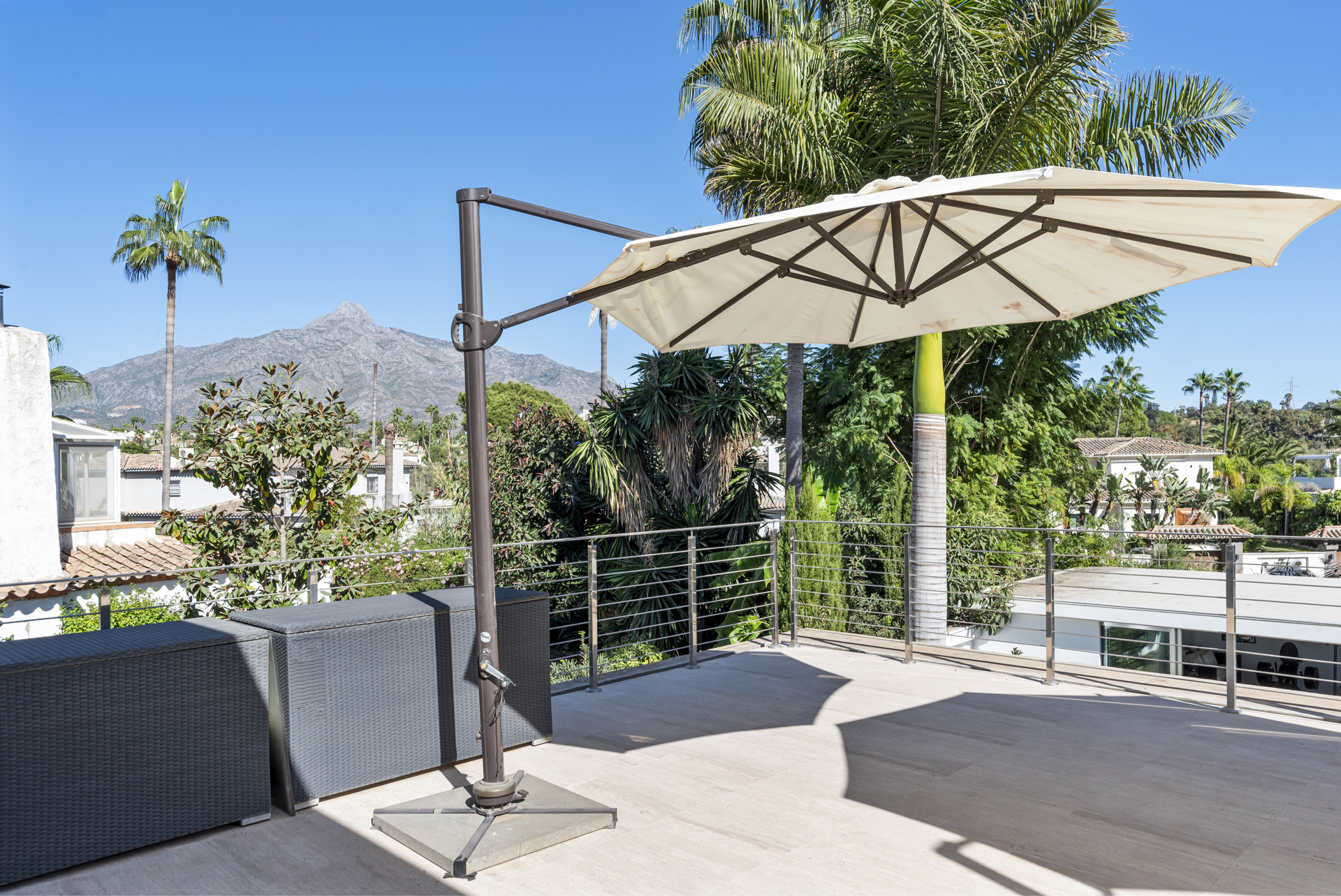 Outdoor terrace with a beige umbrella, dark outdoor storage, steel railing, palm trees, and distant mountains.