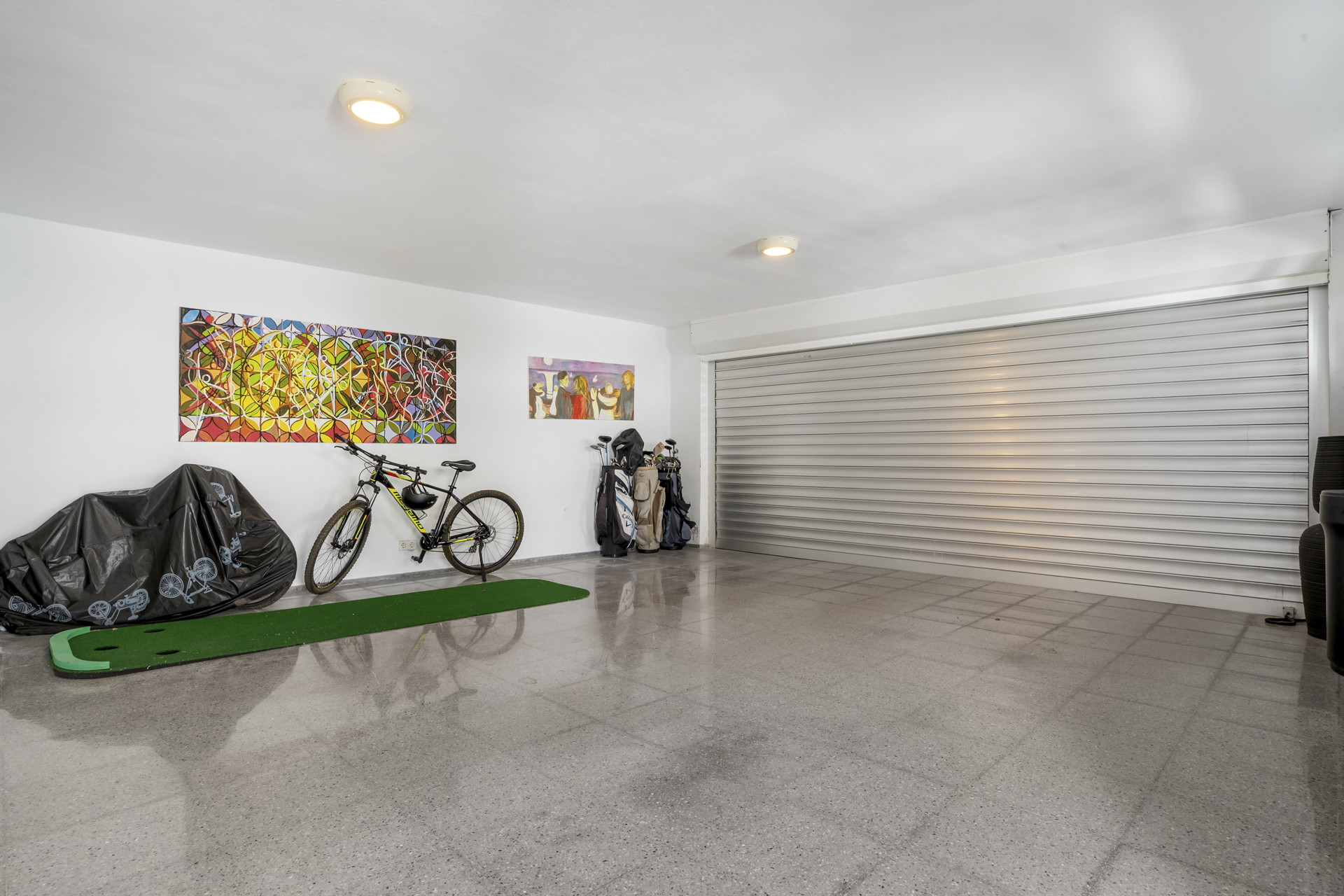 A garage with white walls, a large roller door, concrete floor, colorful art, bicycles, and golf bags.