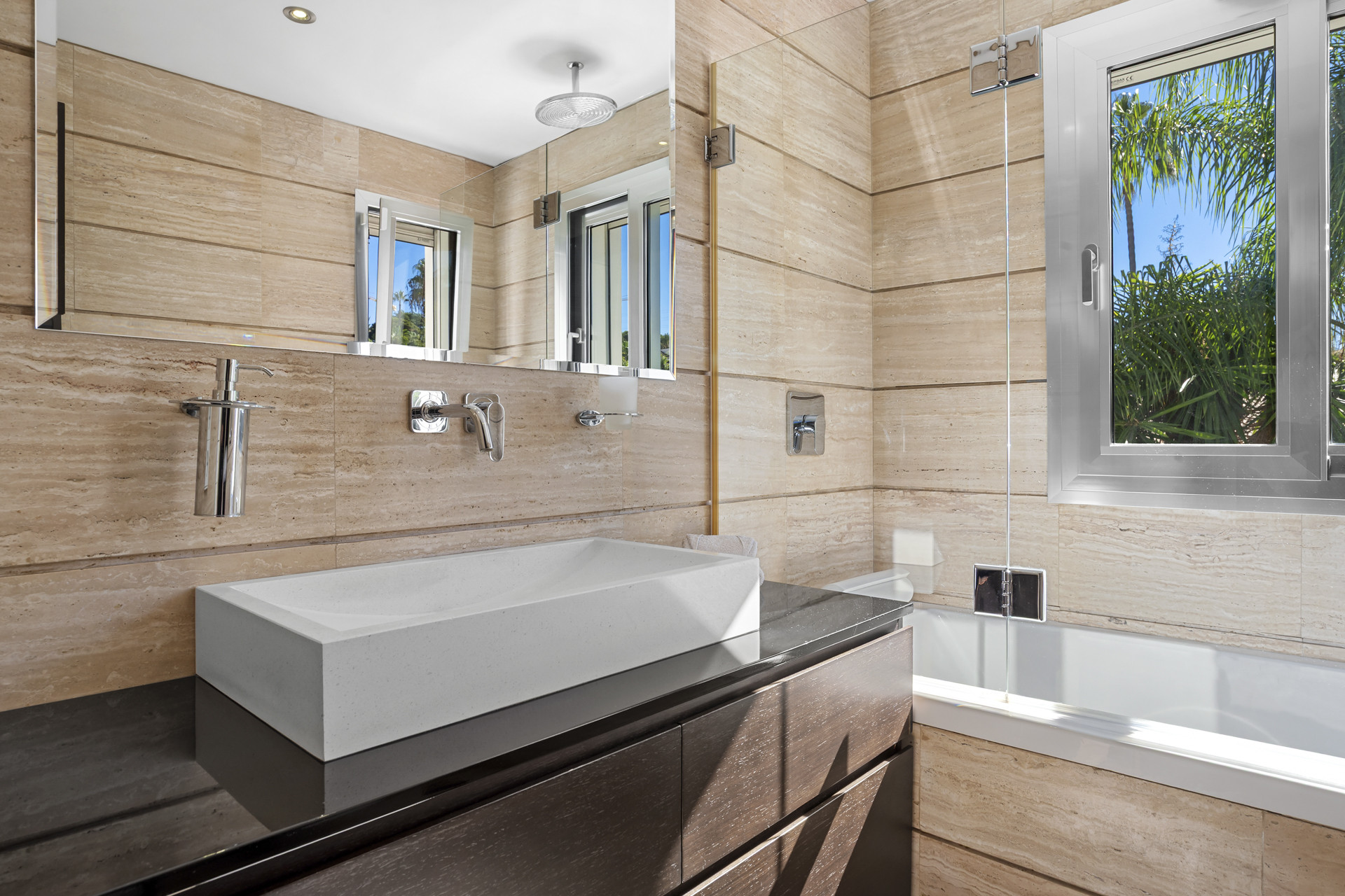 Bathroom with light tiled walls, a white rectangular sink on a dark vanity, a mirror, and a bathtub with a window.
