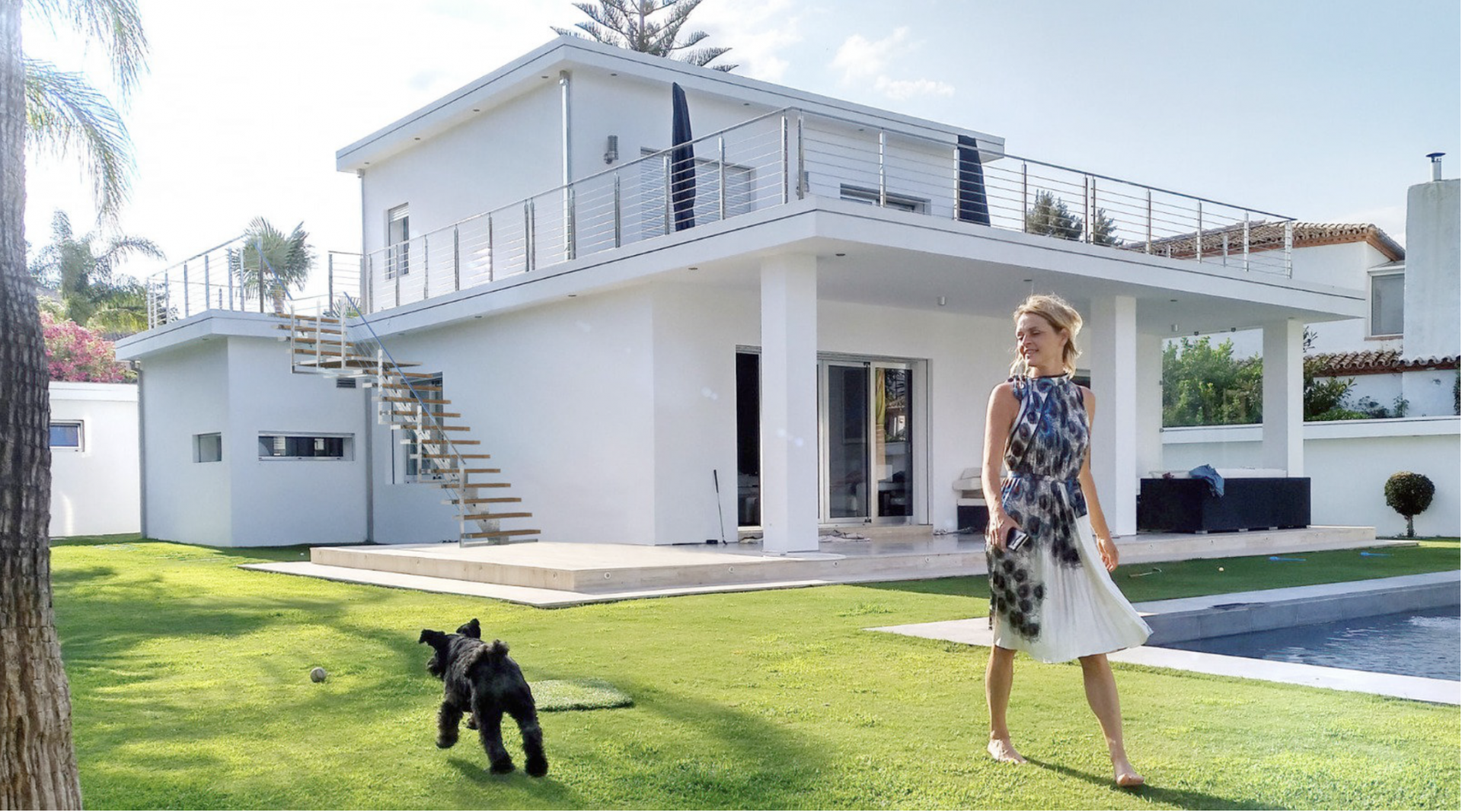 Modern white villa with grass lawn, pool, woman, and dog in the foreground.