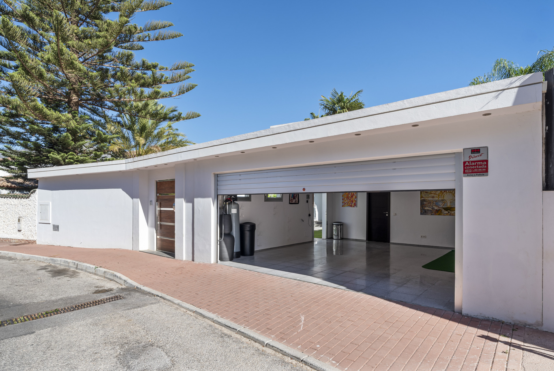 An exterior view of a white garage with an open roller door, a pedestrian entry, and brick paving.