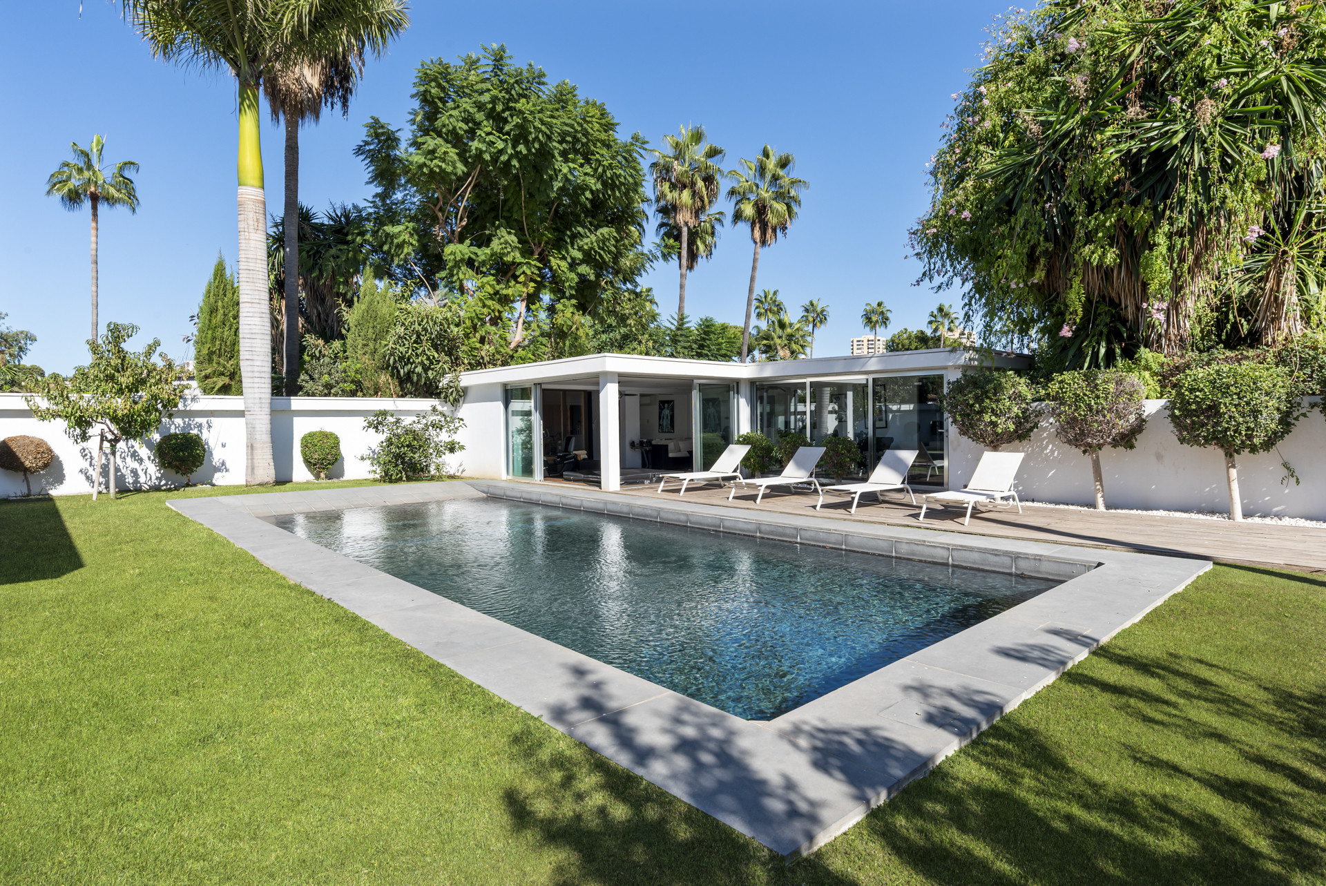 A rectangular swimming pool and a modern pool house with an attached wooden deck, seen from a distance.