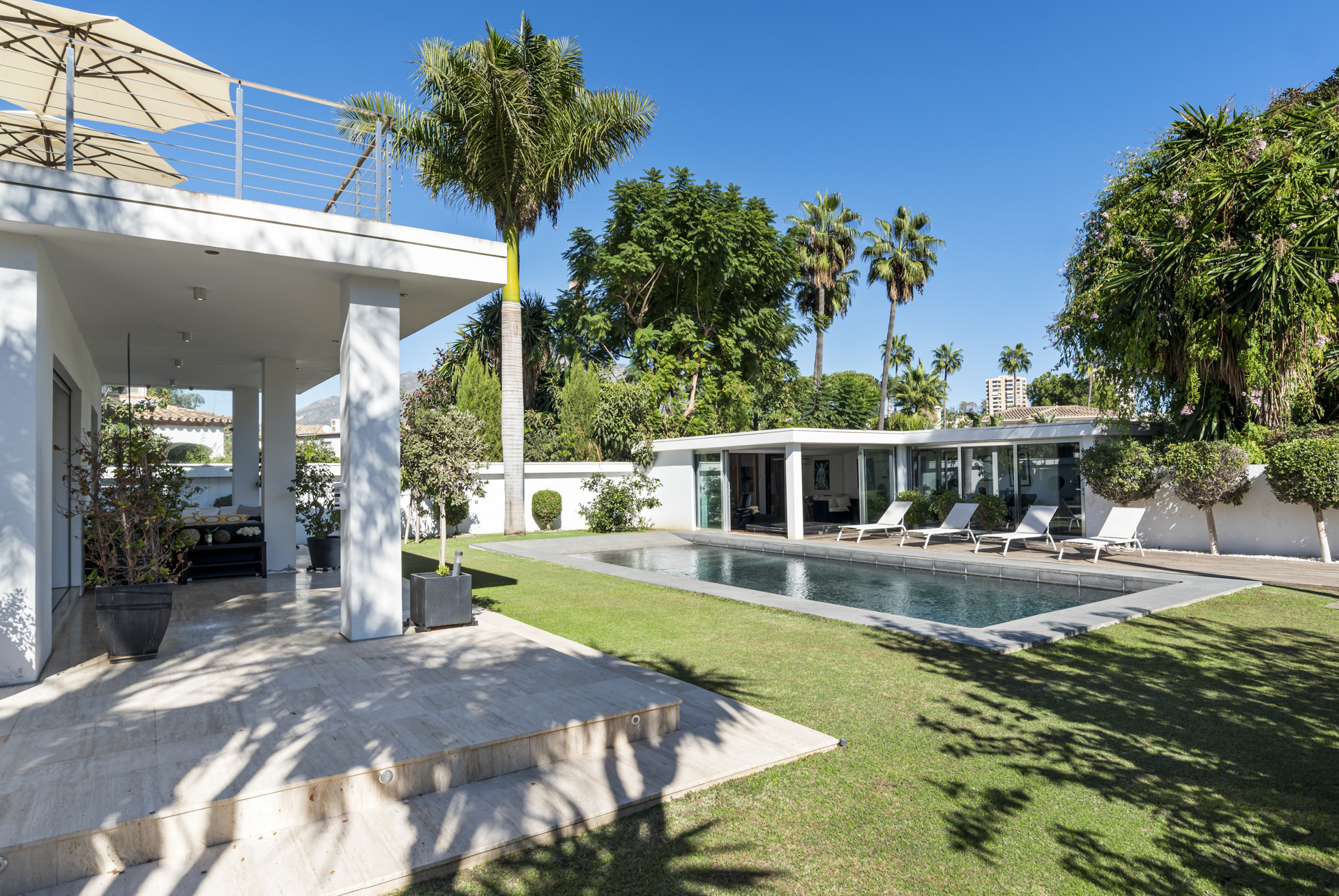 View from a covered patio towards a rectangular swimming pool with four lounge chairs and a pool house.