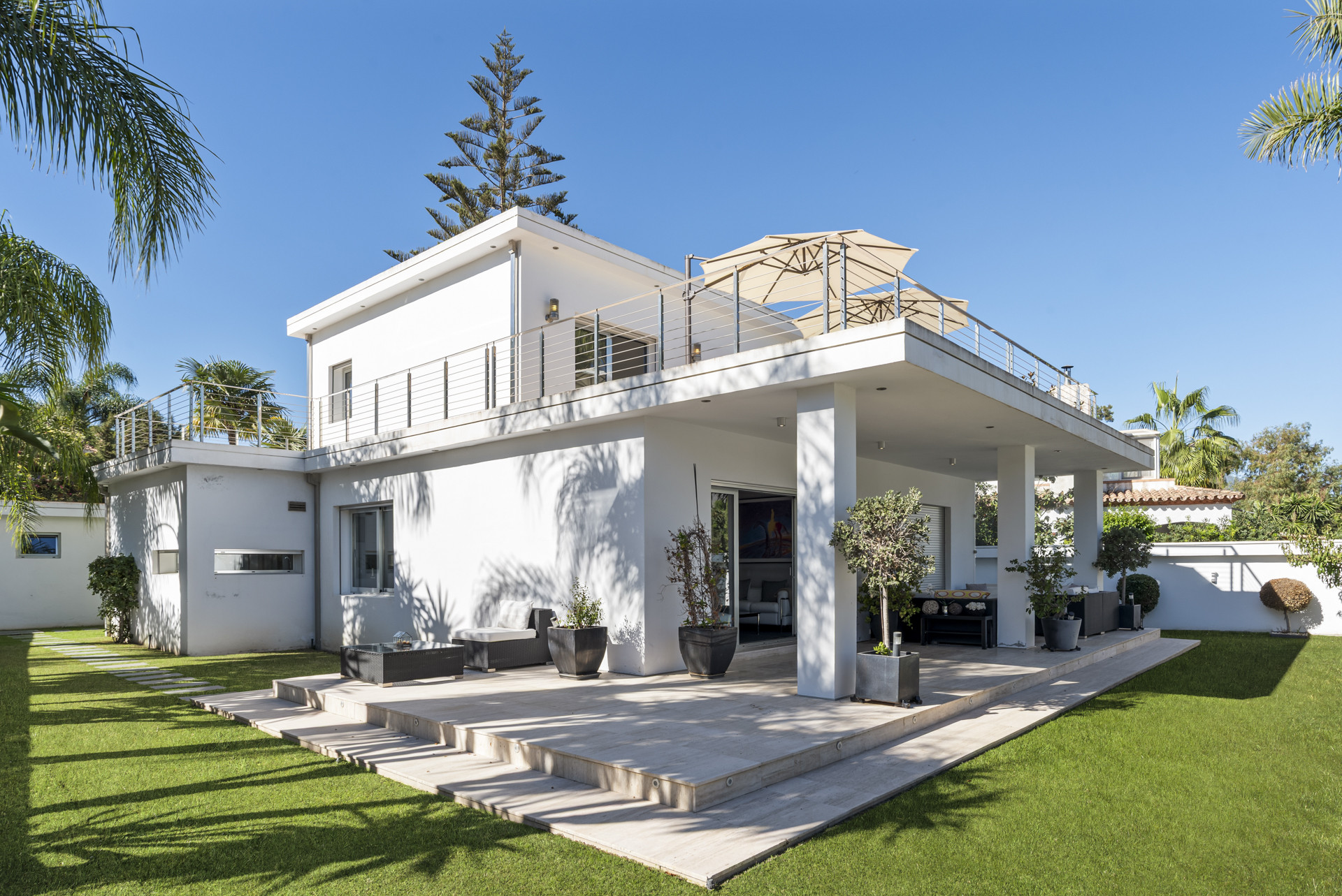 White modern villa exterior showing the back with terraces, steps, and a landscaped green garden.