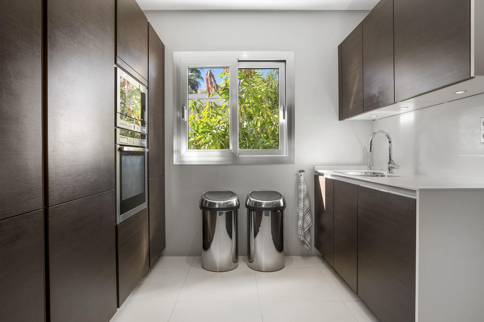 Narrow kitchen aisle with built-in oven, two steel bins, and a window overlooking greenery.