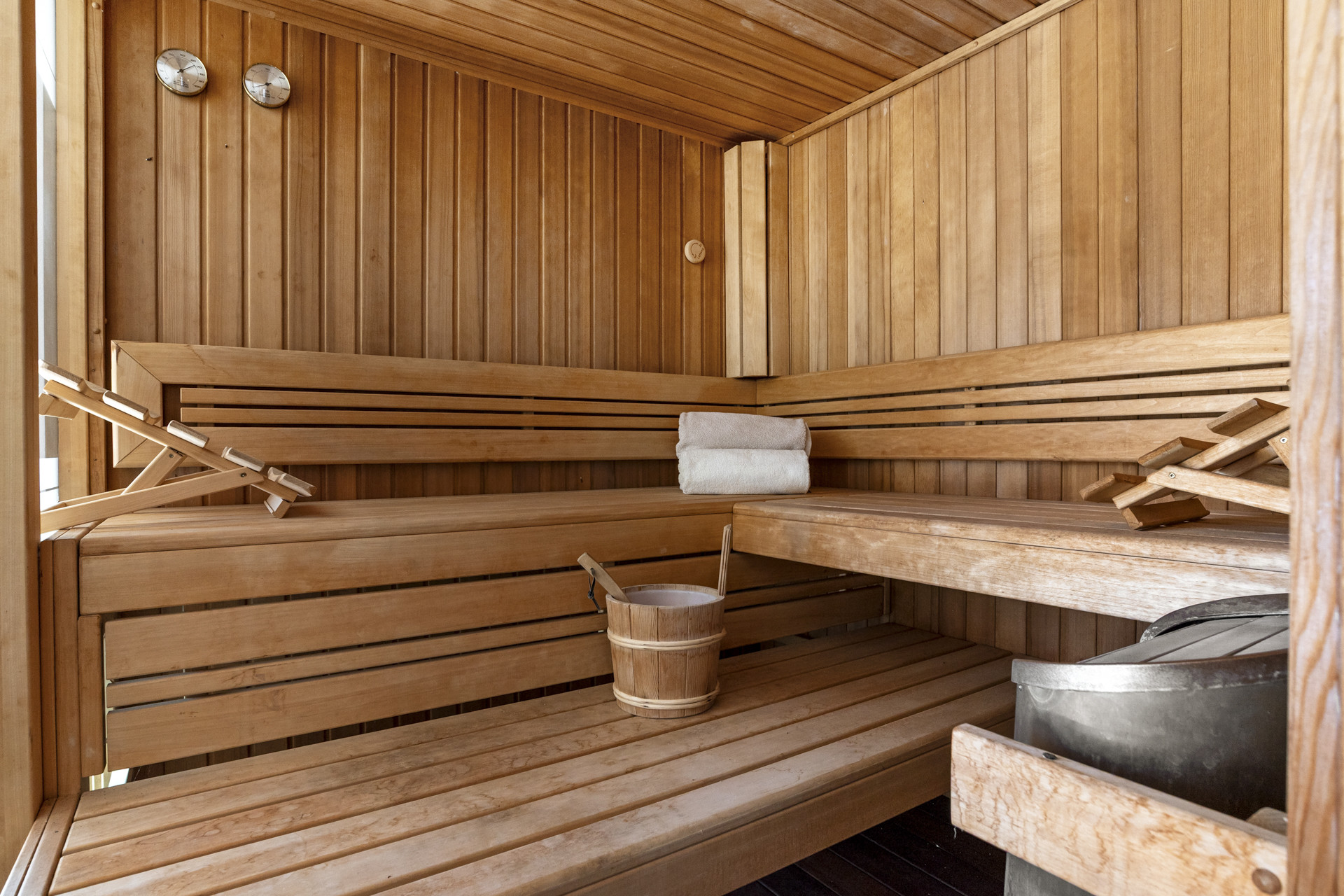 Wooden sauna room with multiple benches, a bucket, and white rolled towels.