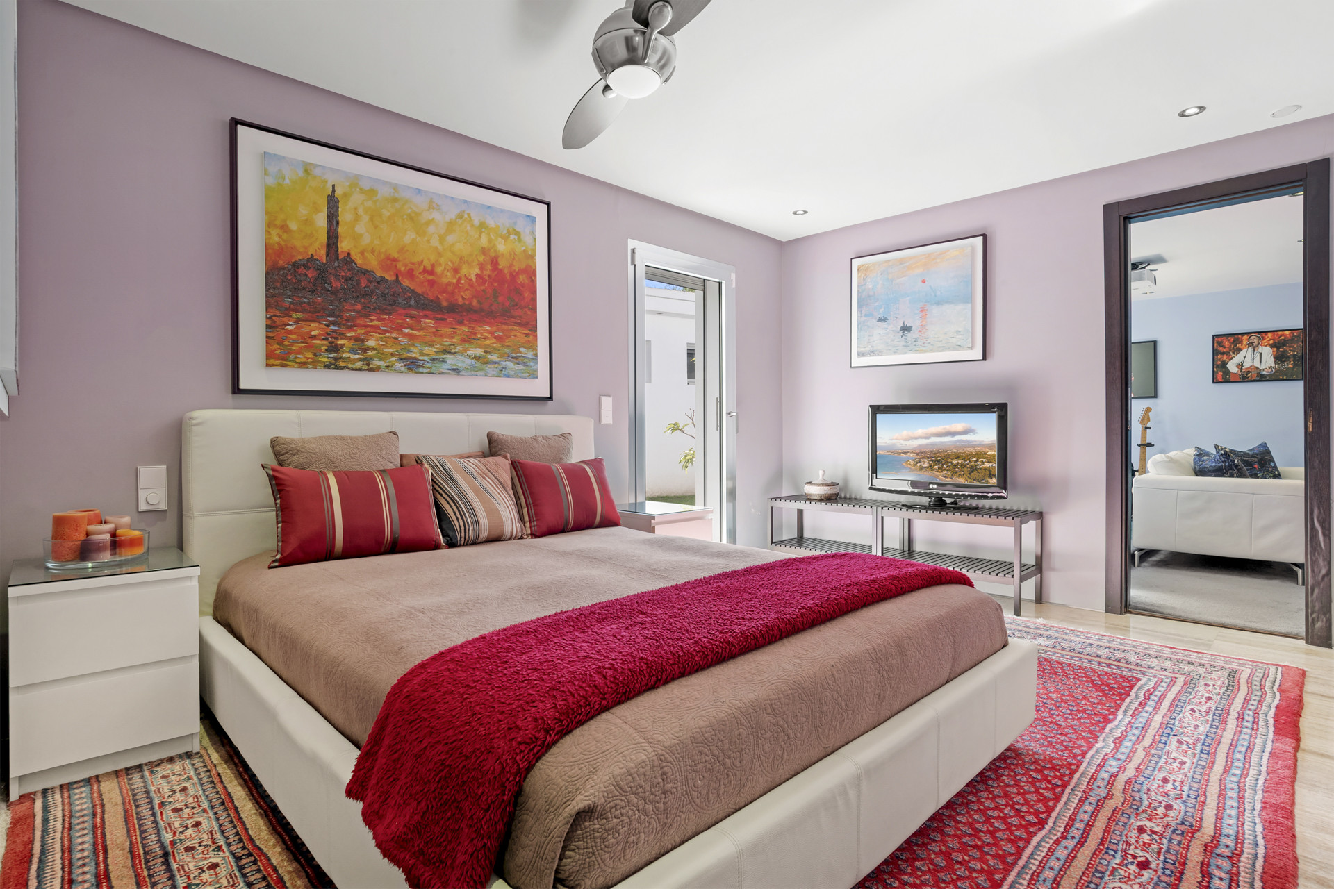 Bedroom with a large bed, purple walls, a red patterned rug, and artwork above the bed.