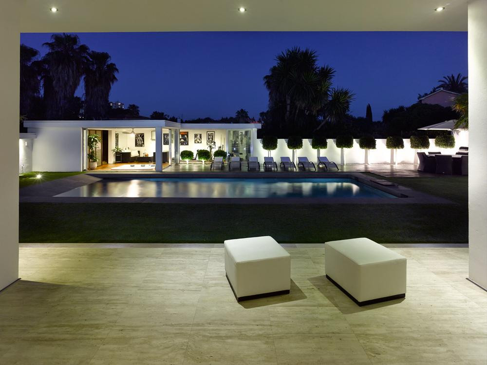 View of a modern white villa and swimming pool at dusk, with outdoor furniture and palm trees.