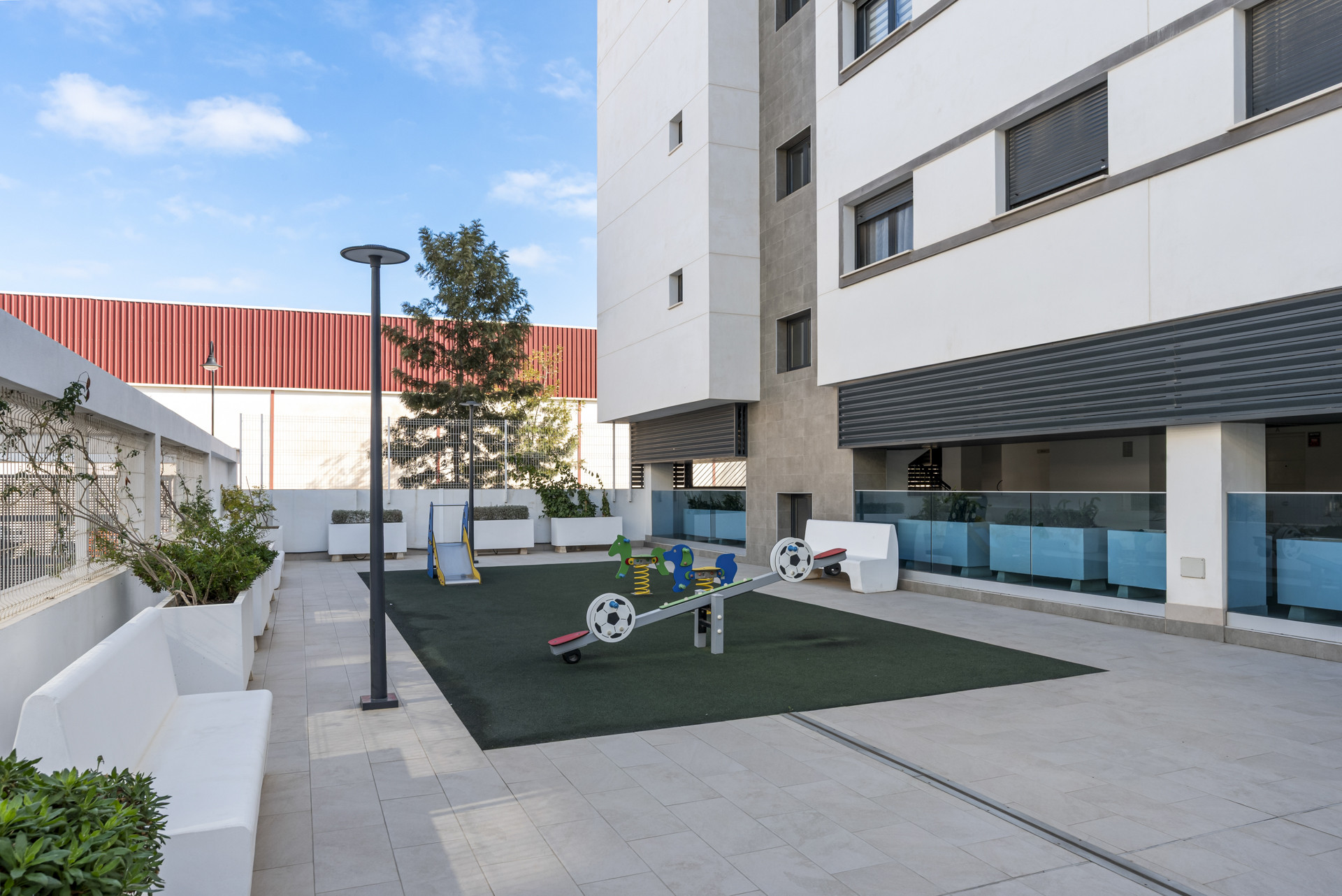 Ground level view of a small children's playground with a seesaw and slide, next to a modern building facade.