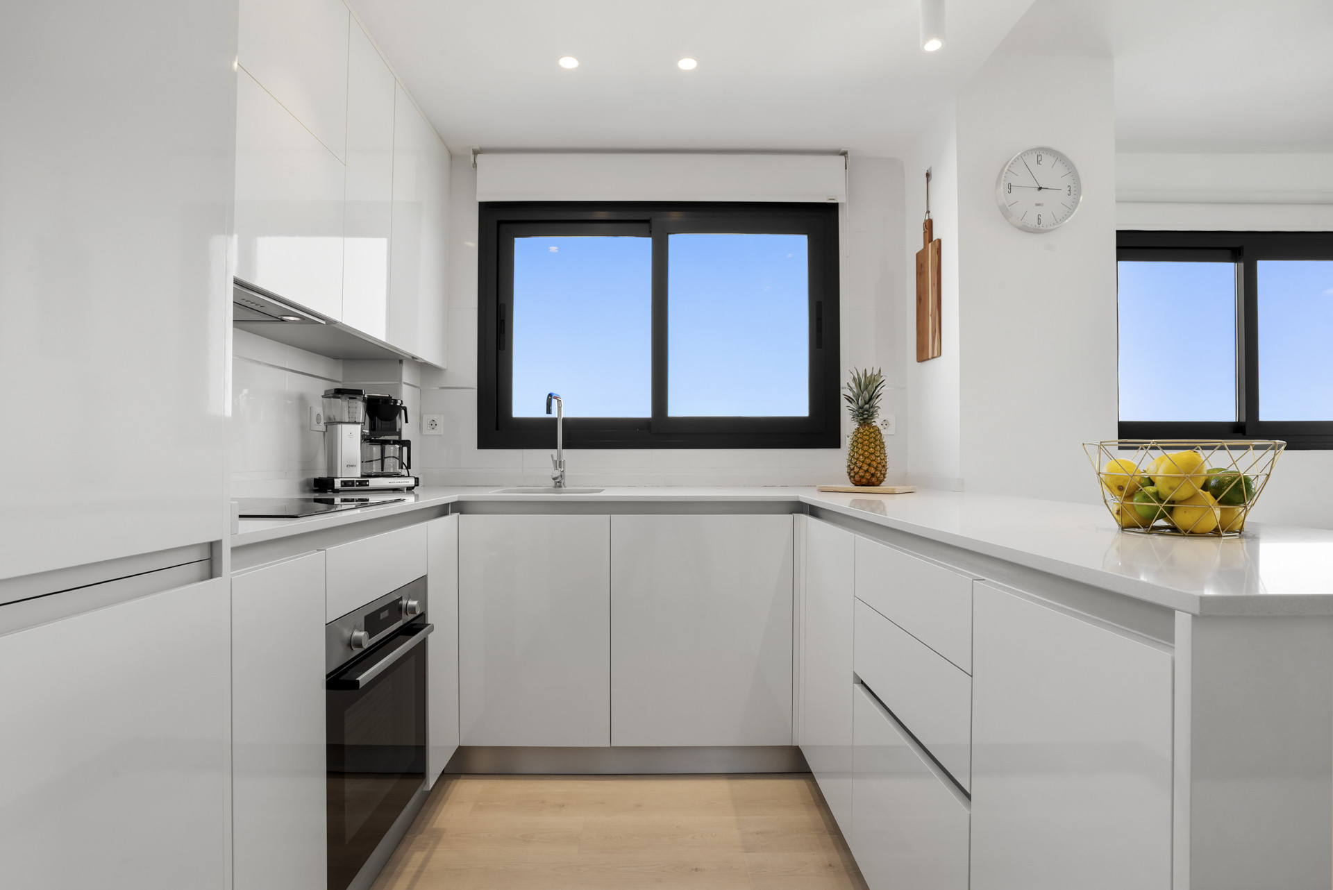 Modern white U-shaped kitchen with black windows, integrated appliances, and a fruit bowl.