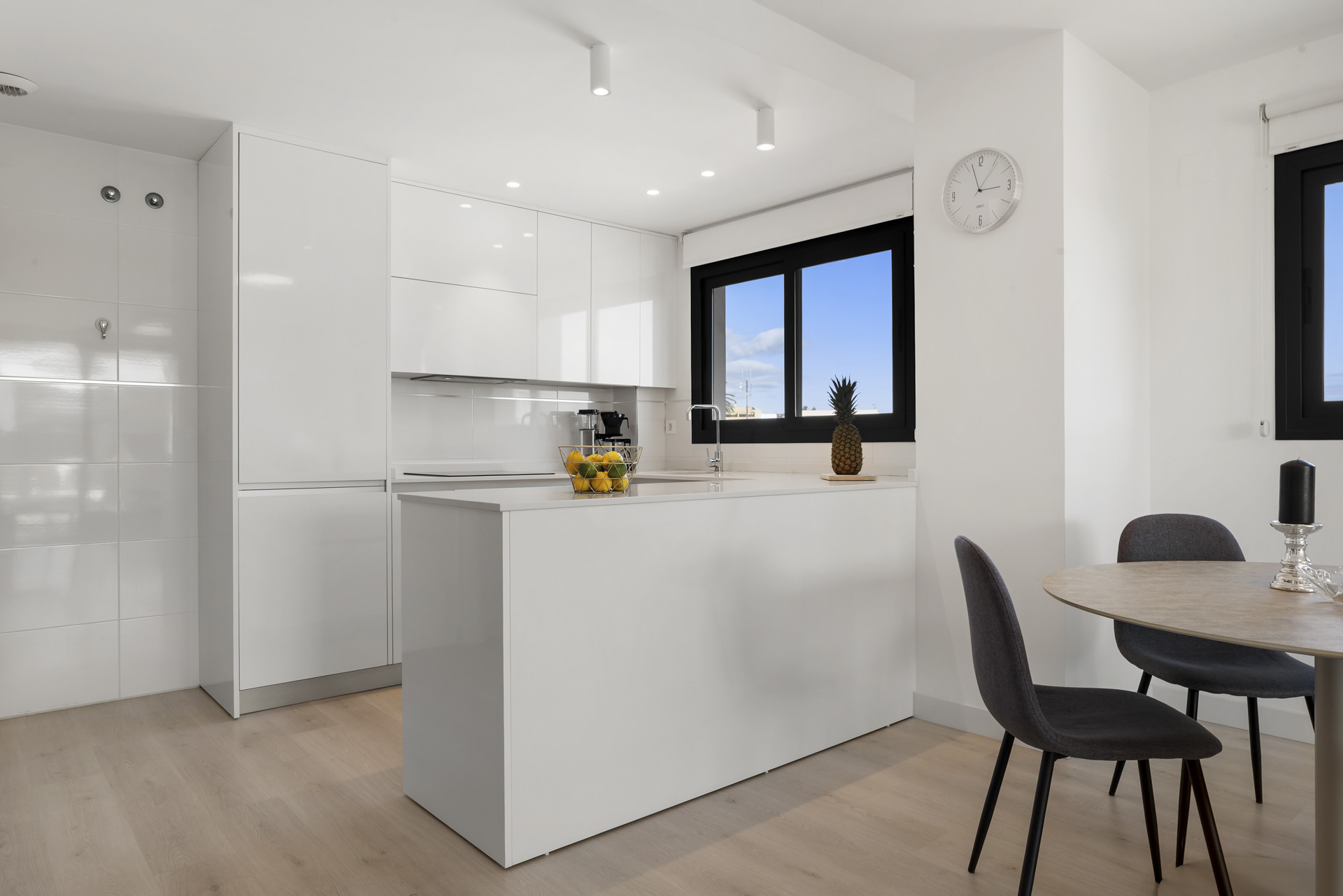 Modern white kitchen with island, wood floor, dining area, and large window overlooking the sky.